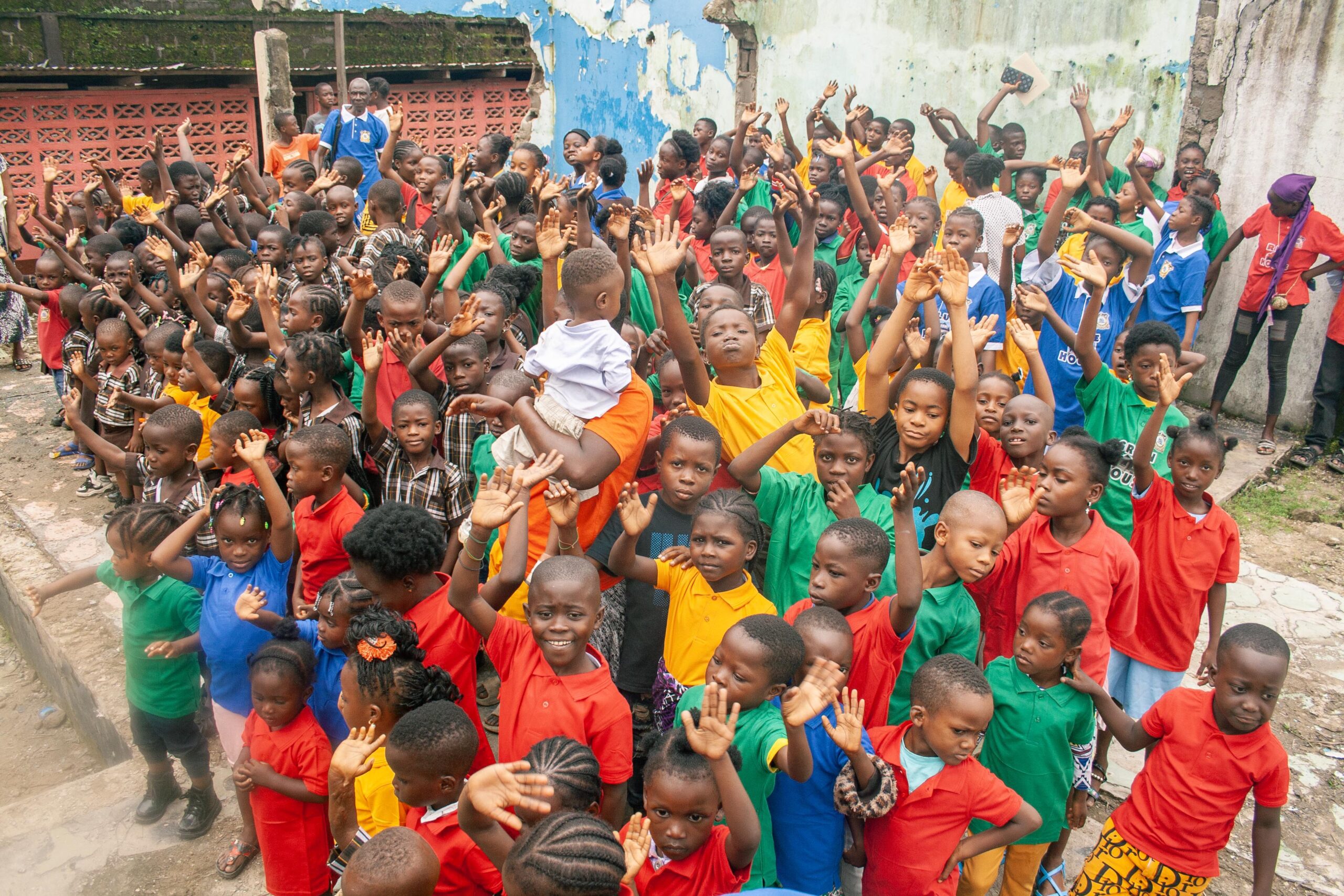 A vibrant group of children wearing colorful shirts enthusiastically raising their hands in an outdoor setting, showcasing a sense of community and joy.