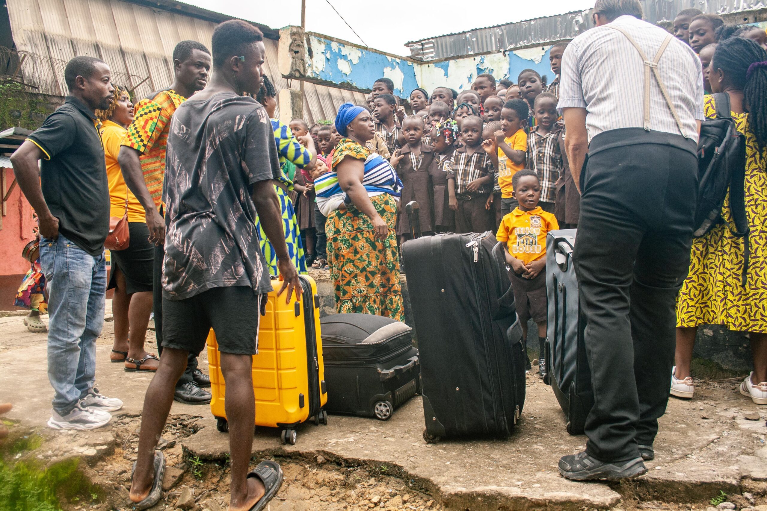 Group of adults and children gathered around luggage in a vibrant community setting, showcasing a mix of traditional and modern attire, with expressions of curiosity and excitement.