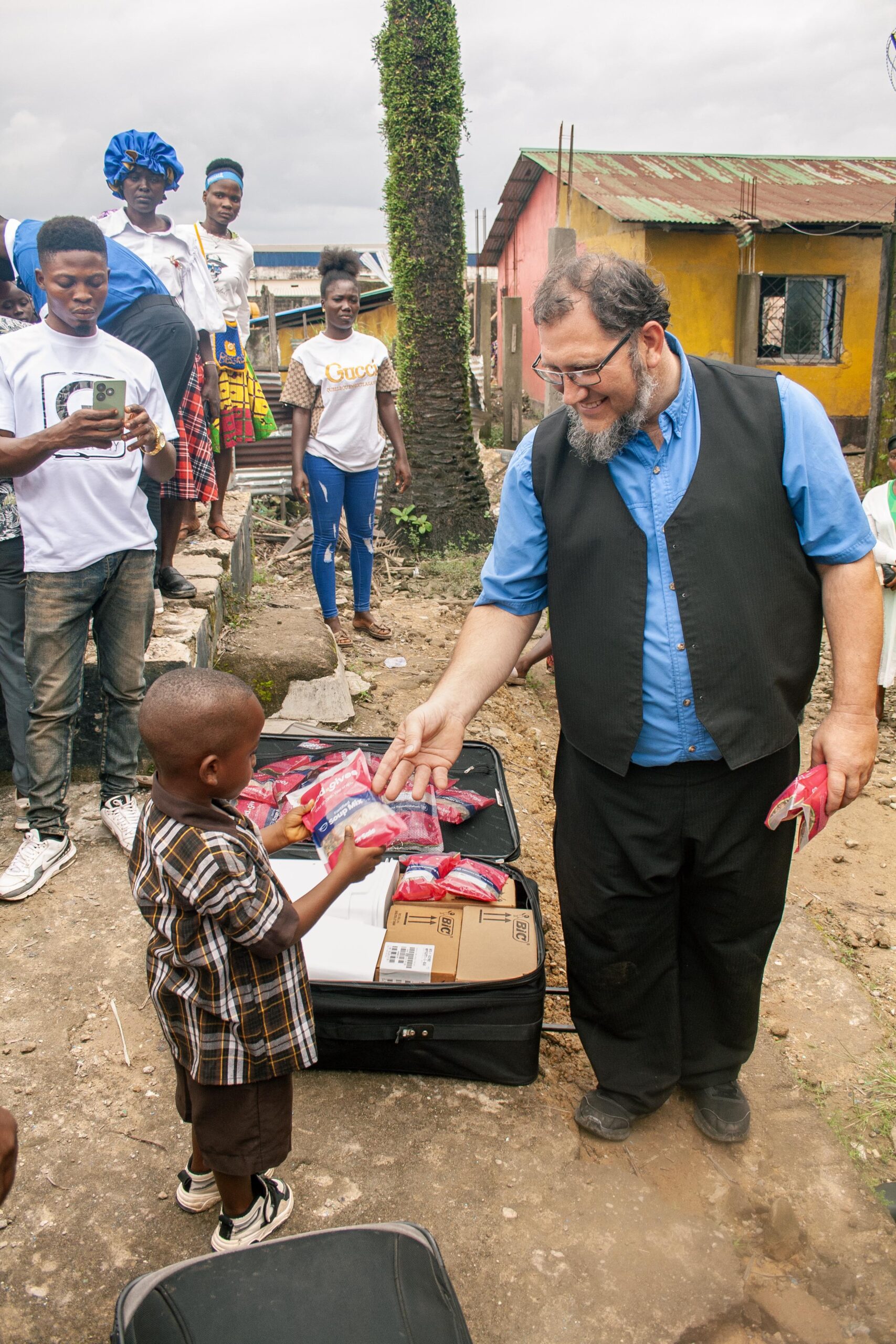 A man in a black vest hands a red package to a young boy in a checkered shirt during a community outreach event in a vibrant neighborhood. In the background, a group of onlookers watches, capturing the moment on their phones. The scene showcases community engagement and the spirit of giving.