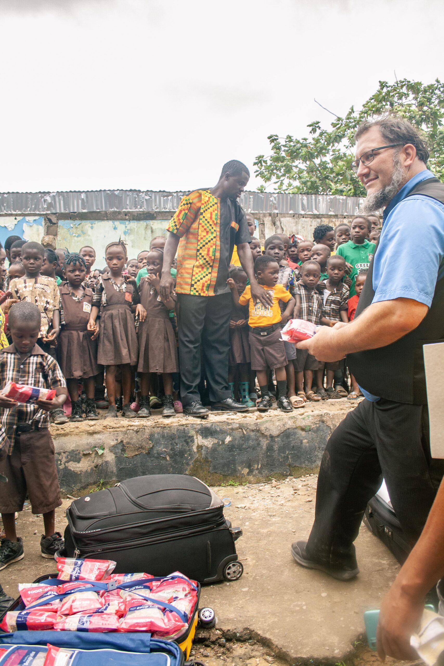 Children in school uniforms gather around two adults distributing supplies in a community setting. The scene depicts a charitable event focused on education and support for local youth, showcasing the importance of community engagement.
