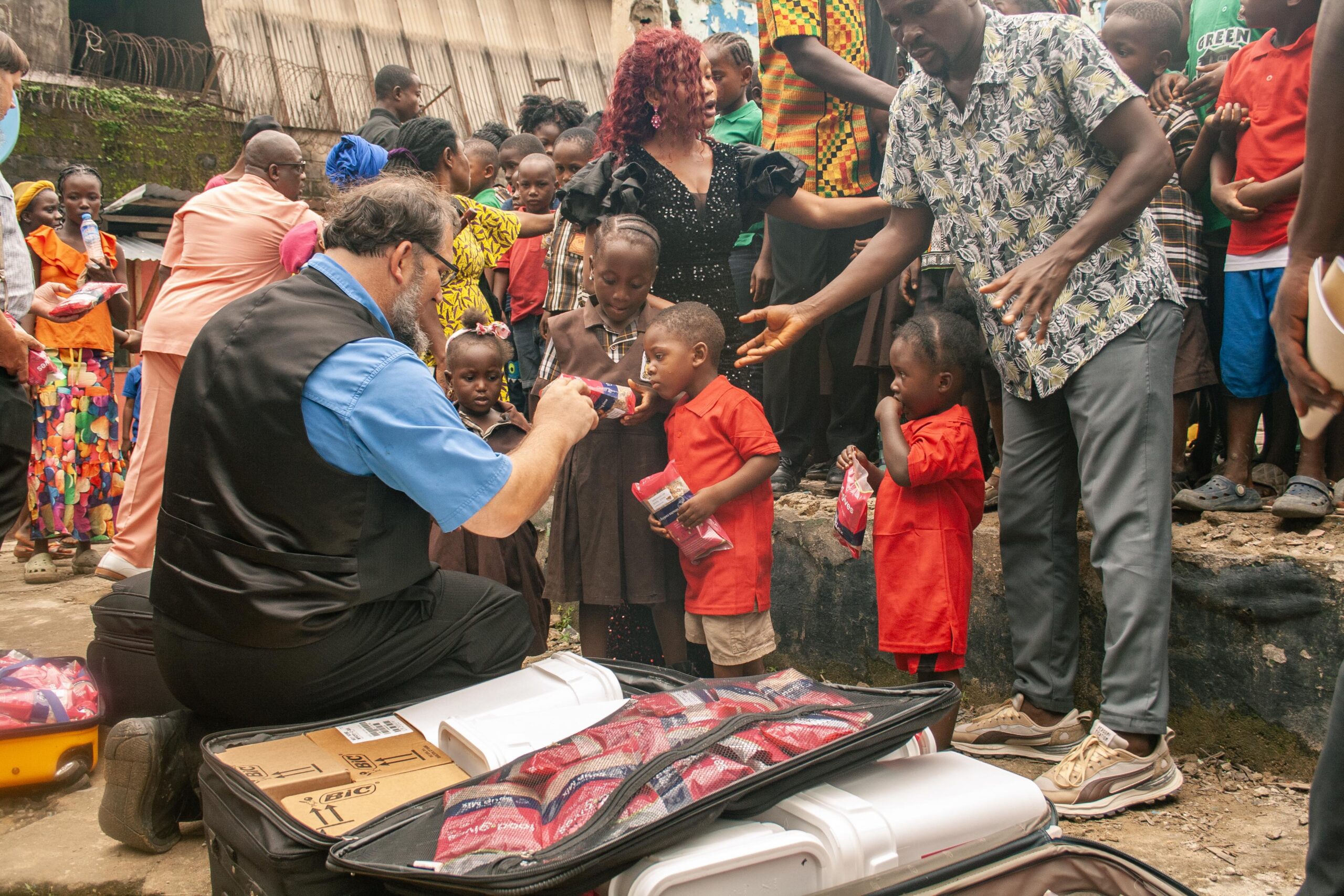 Children receiving aid in a community outreach event, with an adult distributing items from a suitcase. The scene is lively, featuring a diverse group of people, including children in colorful clothing and adults engaging with them.
