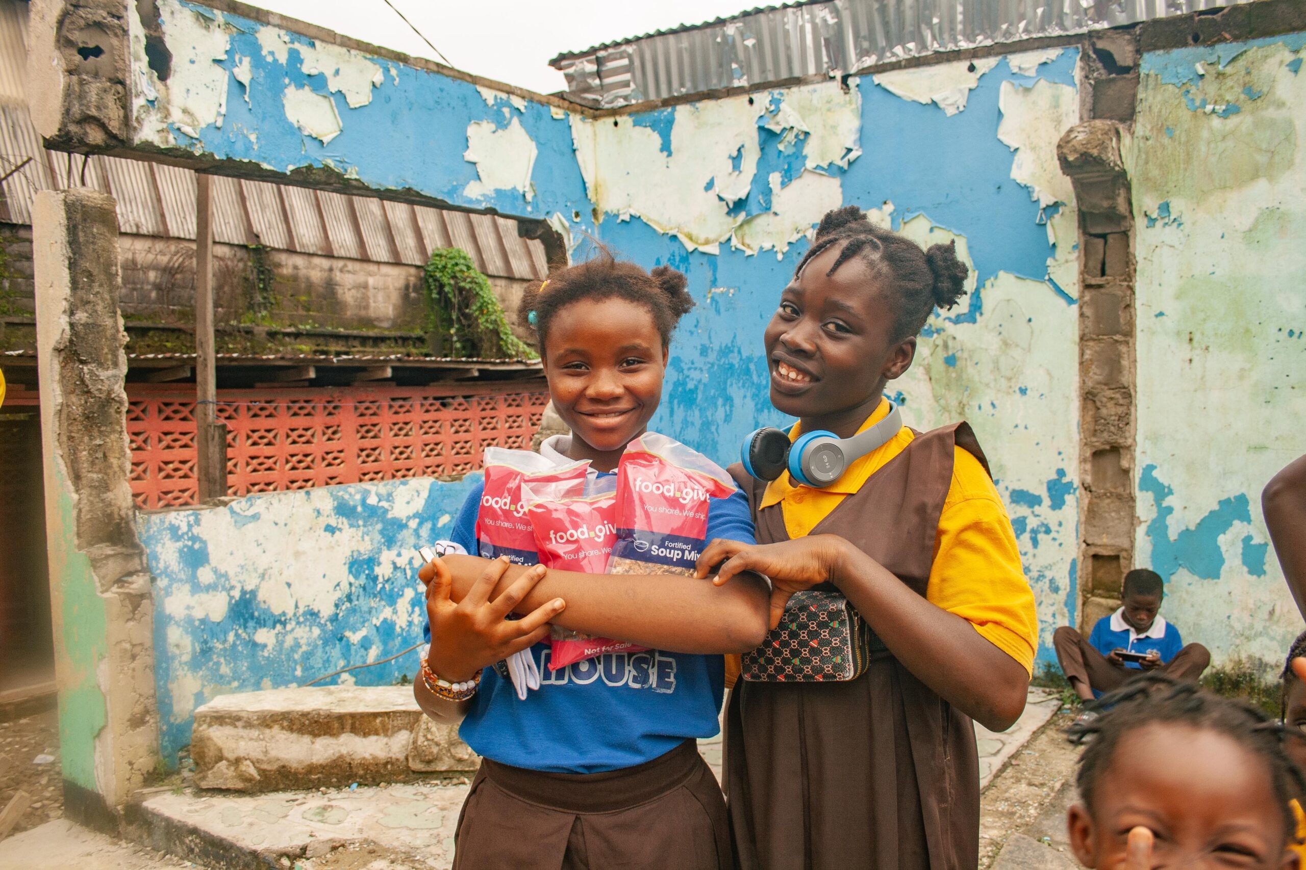 Two smiling girls hold packages of food donations in front of a colorful, weathered wall in a community setting. The girls are dressed in school uniforms, showcasing a sense of joy and community support.