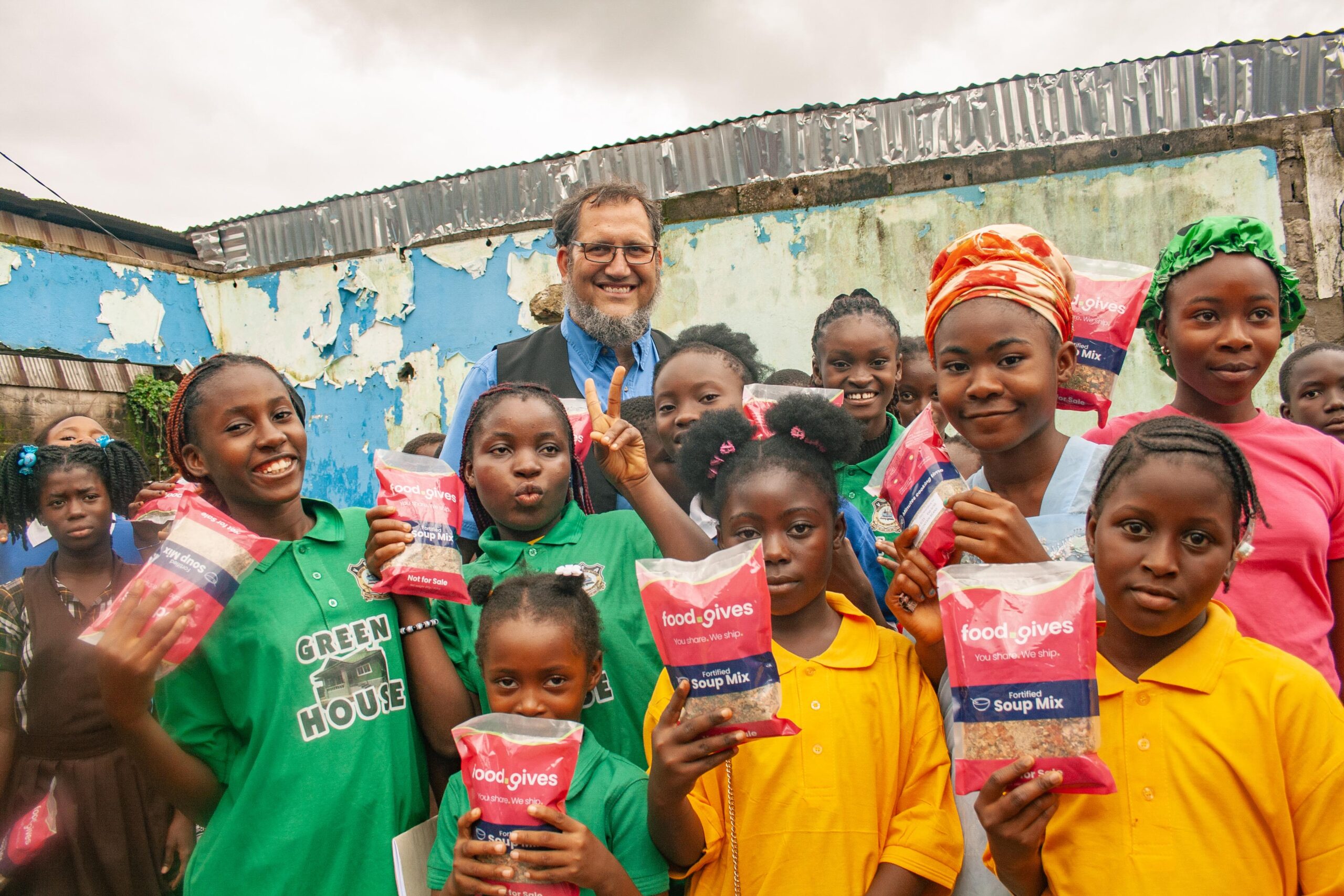 A diverse group of children and an adult man smile and pose for the camera while holding packages of fortified soup mix. The scene takes place against a backdrop of a weathered blue wall, highlighting a community food distribution initiative. The children are wearing colorful shirts, and the atmosphere is joyful and supportive, reflecting the spirit of giving and community involvement.