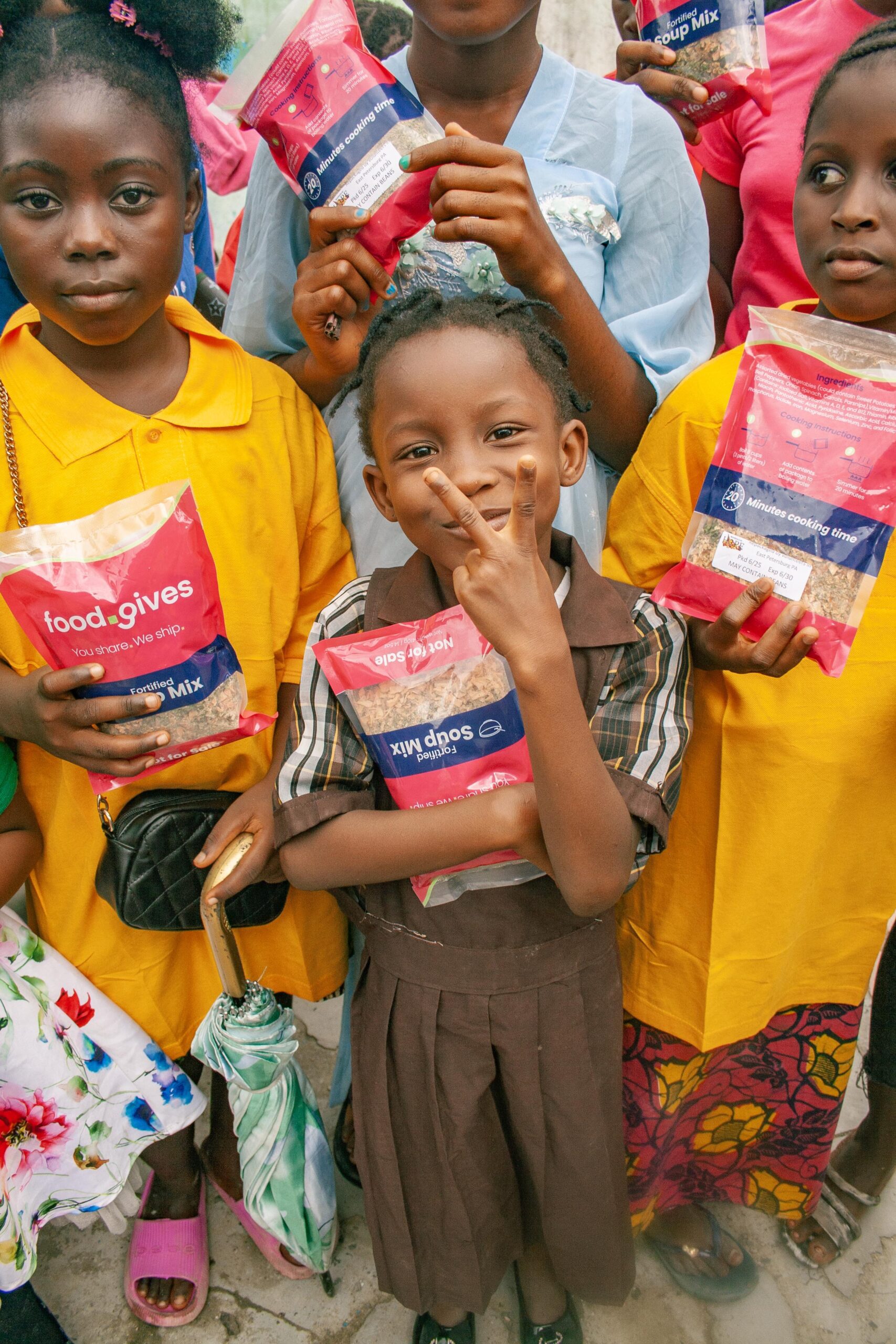 A group of children holding food packages, smiling and showing peace signs, highlighting a community initiative providing nutritious meal options. The children are dressed in colorful clothing, with one girl in a brown dress prominently displaying her food package.