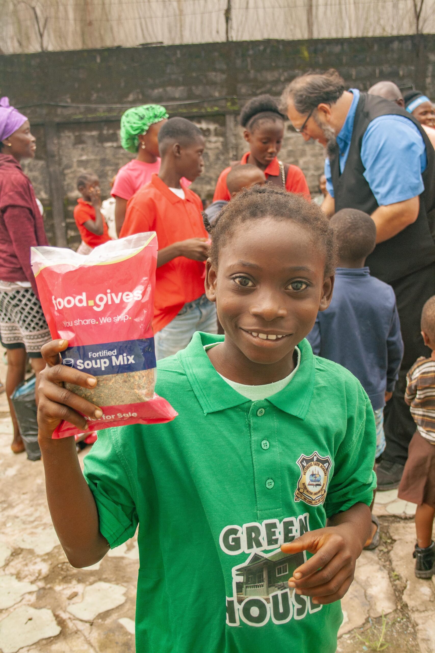 A smiling child in a green shirt holds a packet of fortified soup mix labeled "food.gives" during a community event, surrounded by other children and adults in a vibrant outdoor setting.