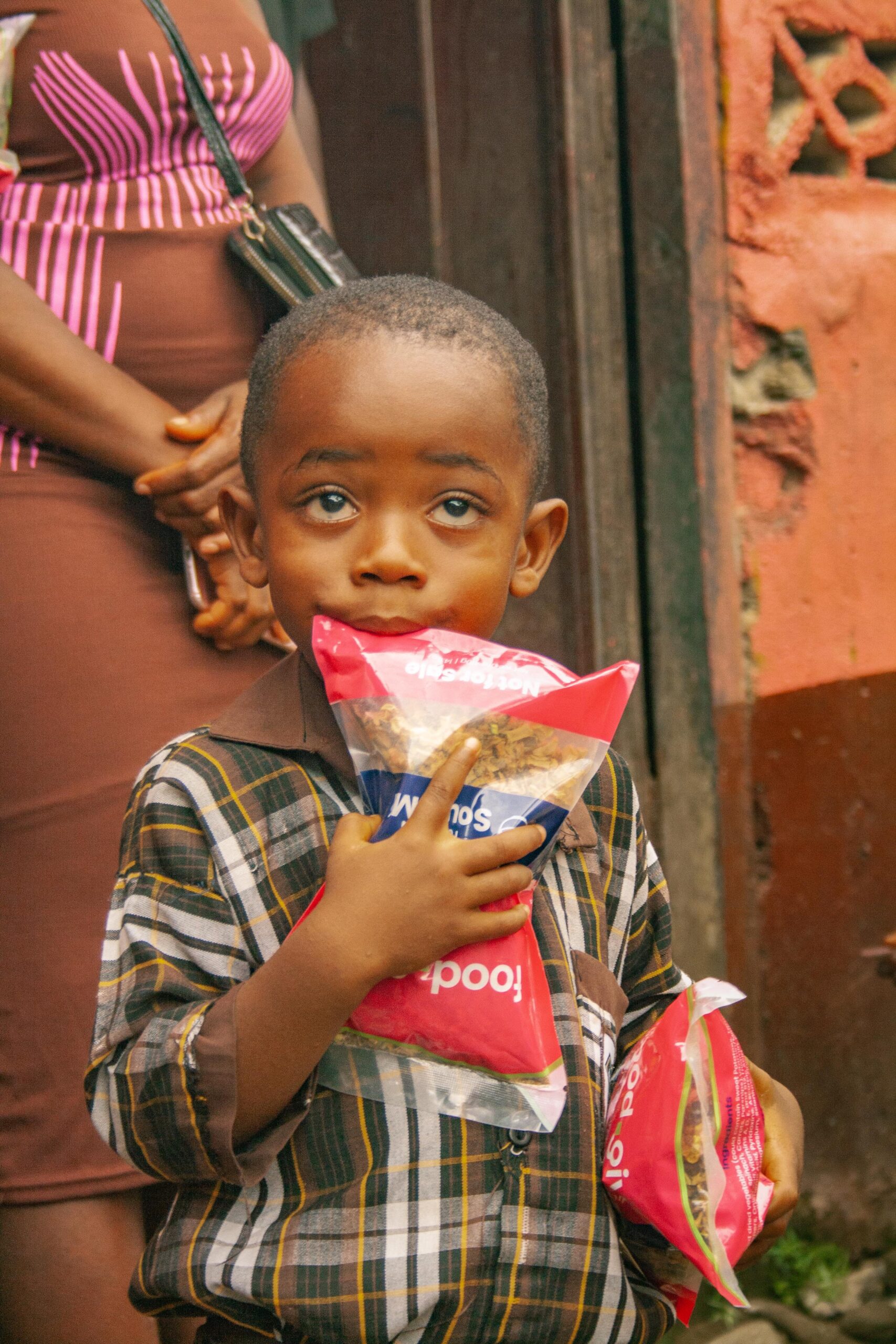 Young boy holding two packets of snacks, looking curiously at the camera, with a blurred background featuring a woman in a striped dress. The scene captures a moment of childhood joy and curiosity in an urban setting.
