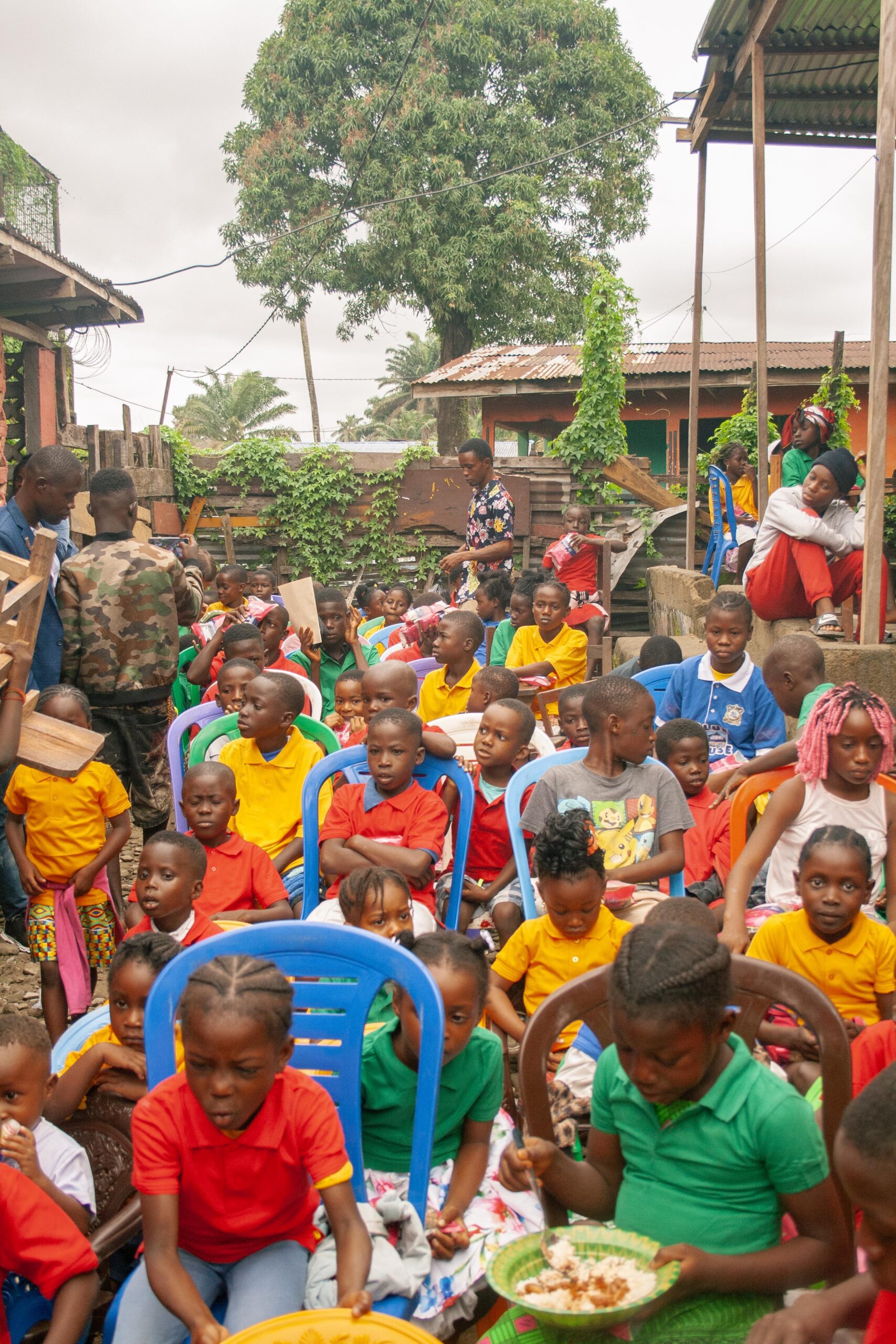 Children gathered in colorful shirts enjoying a community event outdoors, sitting on plastic chairs in a lively atmosphere surrounded by greenery and local structures.