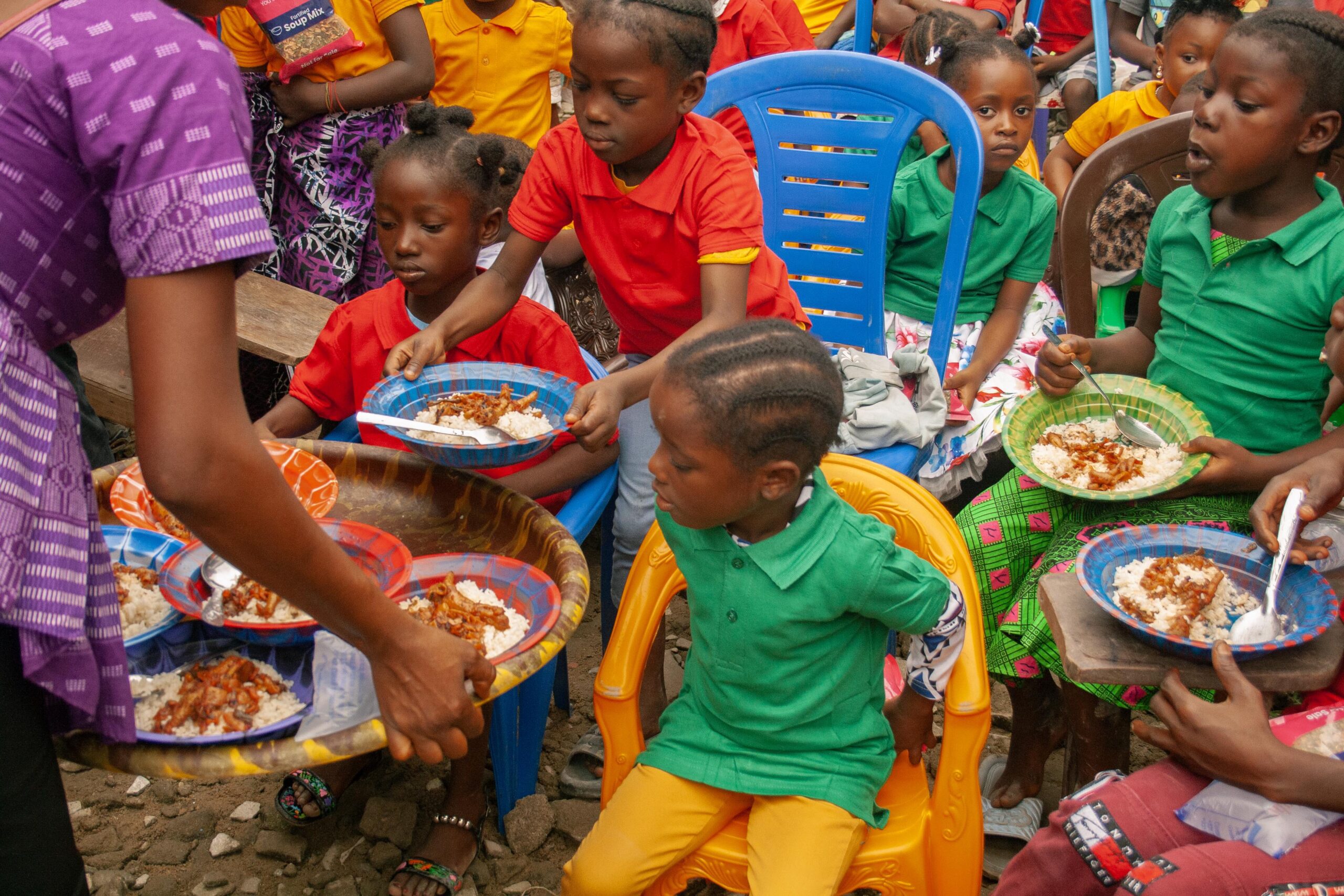 Children sitting in colorful chairs eagerly receiving plates of rice and stew during a community meal event, showcasing a vibrant atmosphere of sharing and joy.