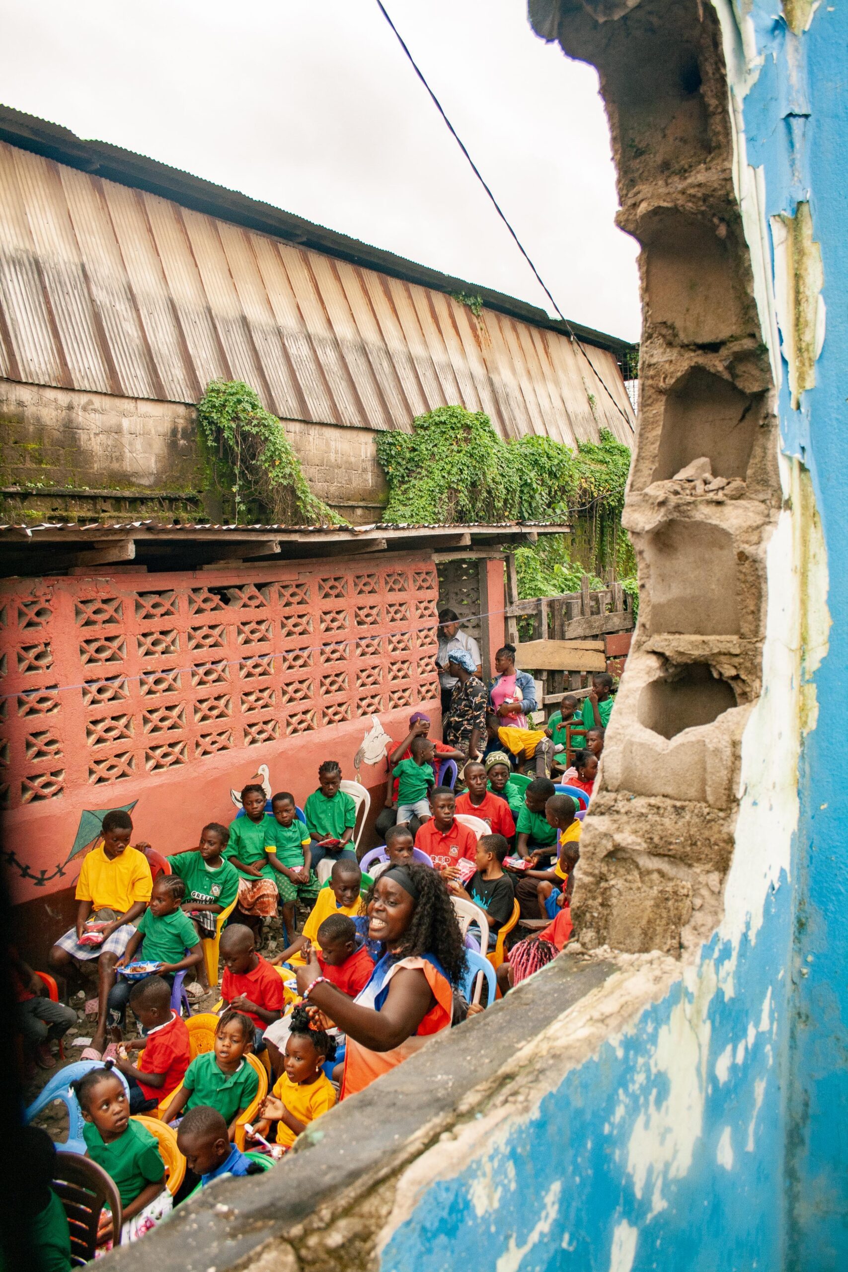 A vibrant community gathering in an outdoor setting, featuring a diverse group of children seated in colorful shirts, engaged in an activity led by an animated adult. The backdrop includes a brick wall and lush greenery, illustrating a lively atmosphere.
