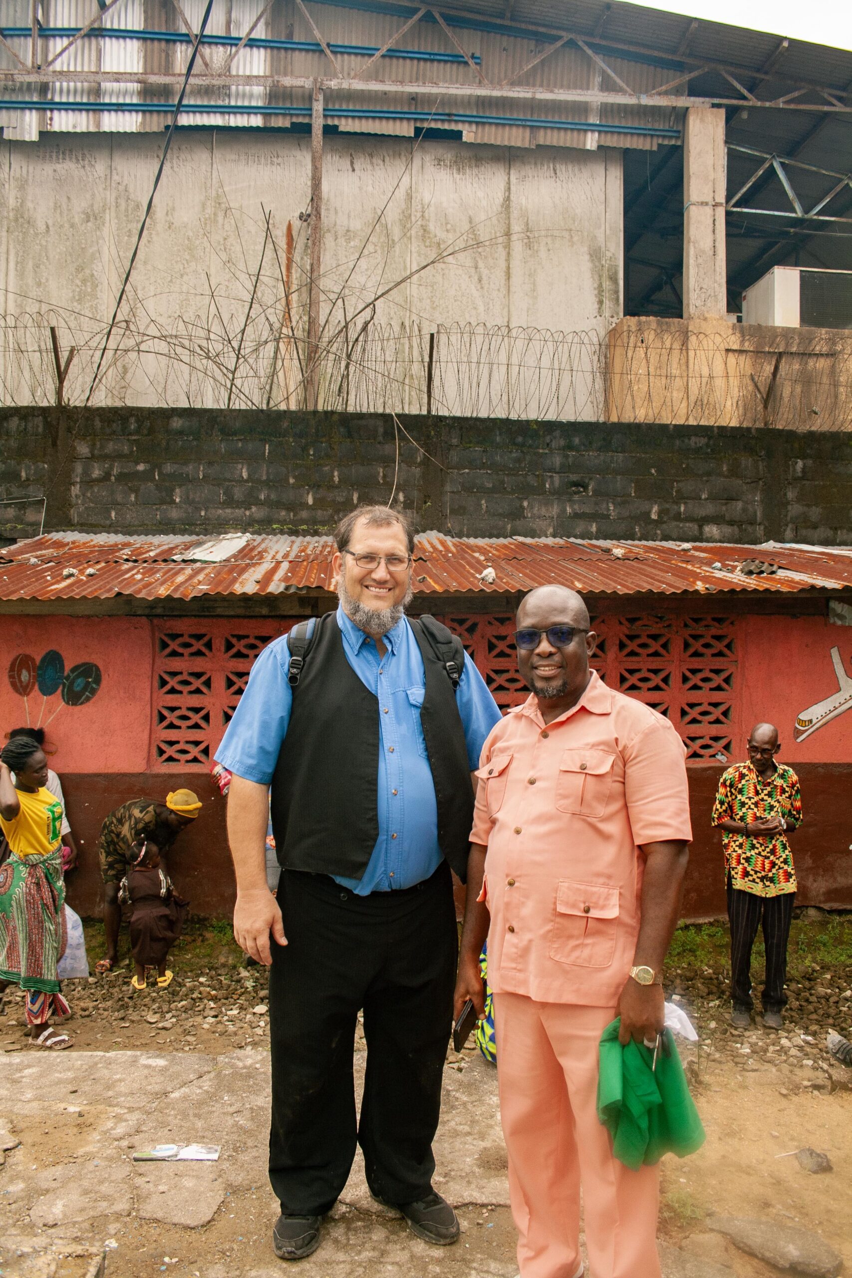 Two men standing together in front of a colorful wall in an urban setting, with a background featuring a corrugated metal structure and barbed wire. The man on the left is wearing a blue shirt and black vest, while the man on the right is dressed in a light pink outfit. People in traditional attire can be seen in the background, highlighting local culture.