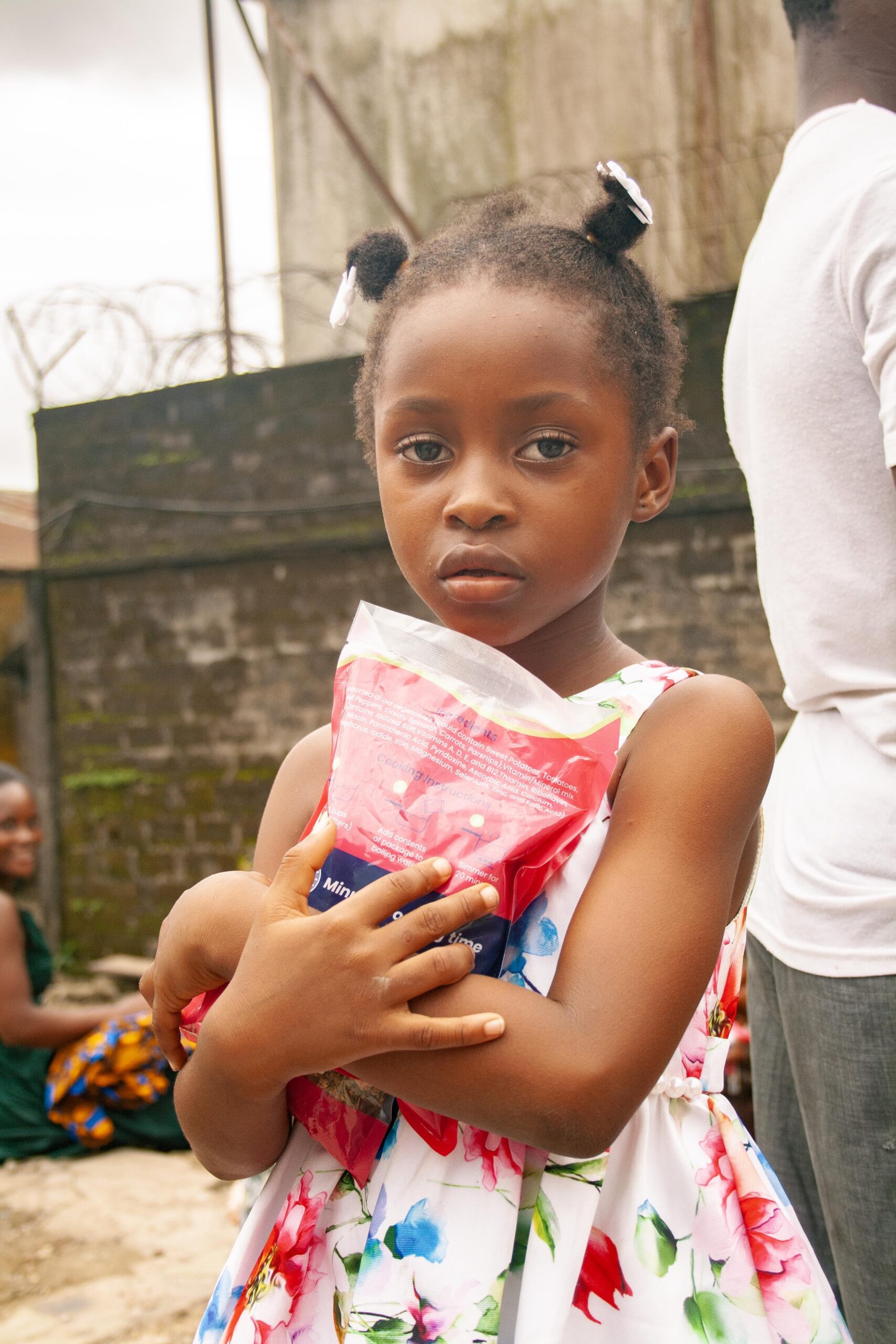 Young girl holding a bag of food, wearing a floral dress, with a serious expression, set against a backdrop of a community environment.