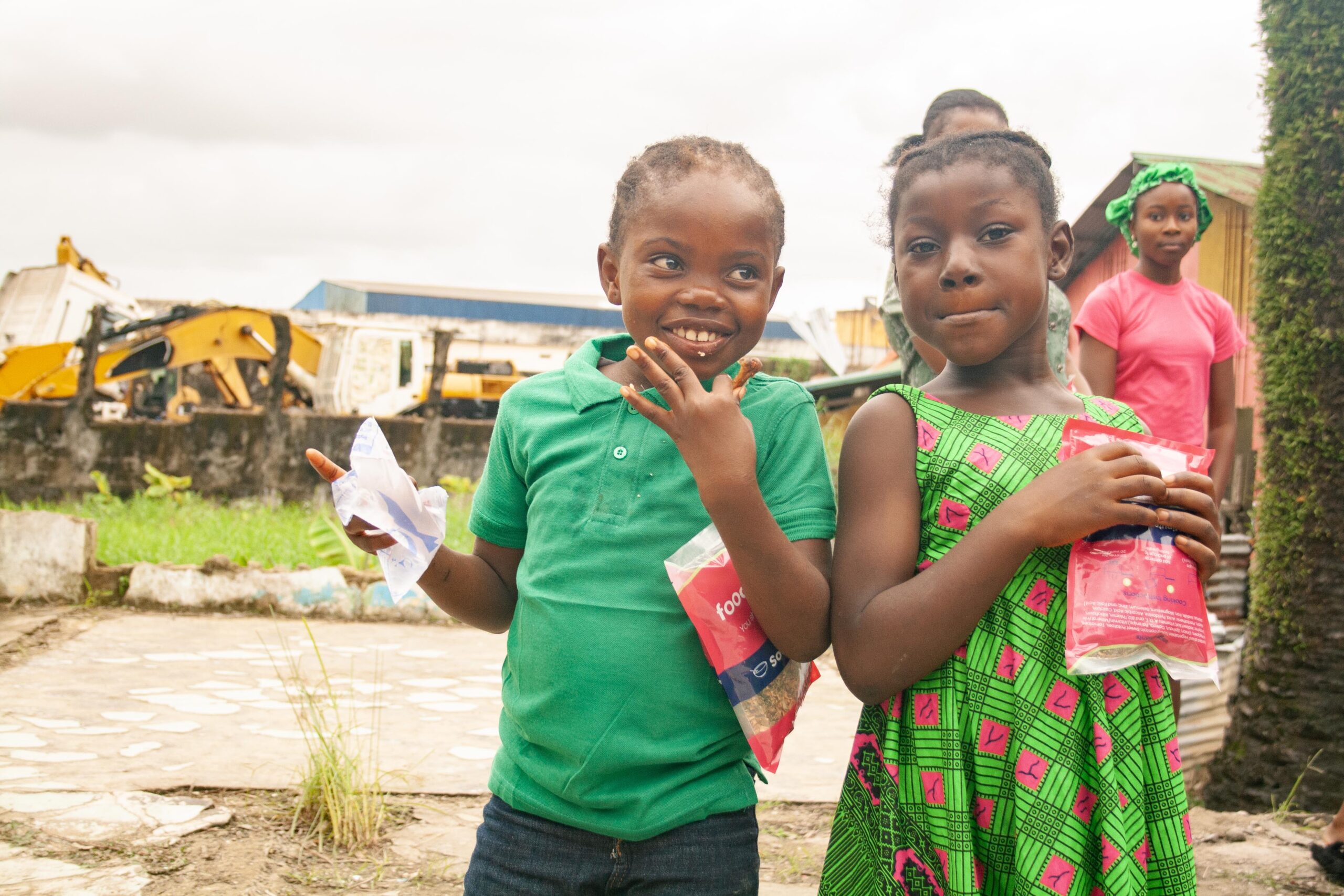 Children smiling and enjoying snacks outdoors, wearing colorful clothing, with a construction site in the background. The image captures a joyful moment of community and childhood.