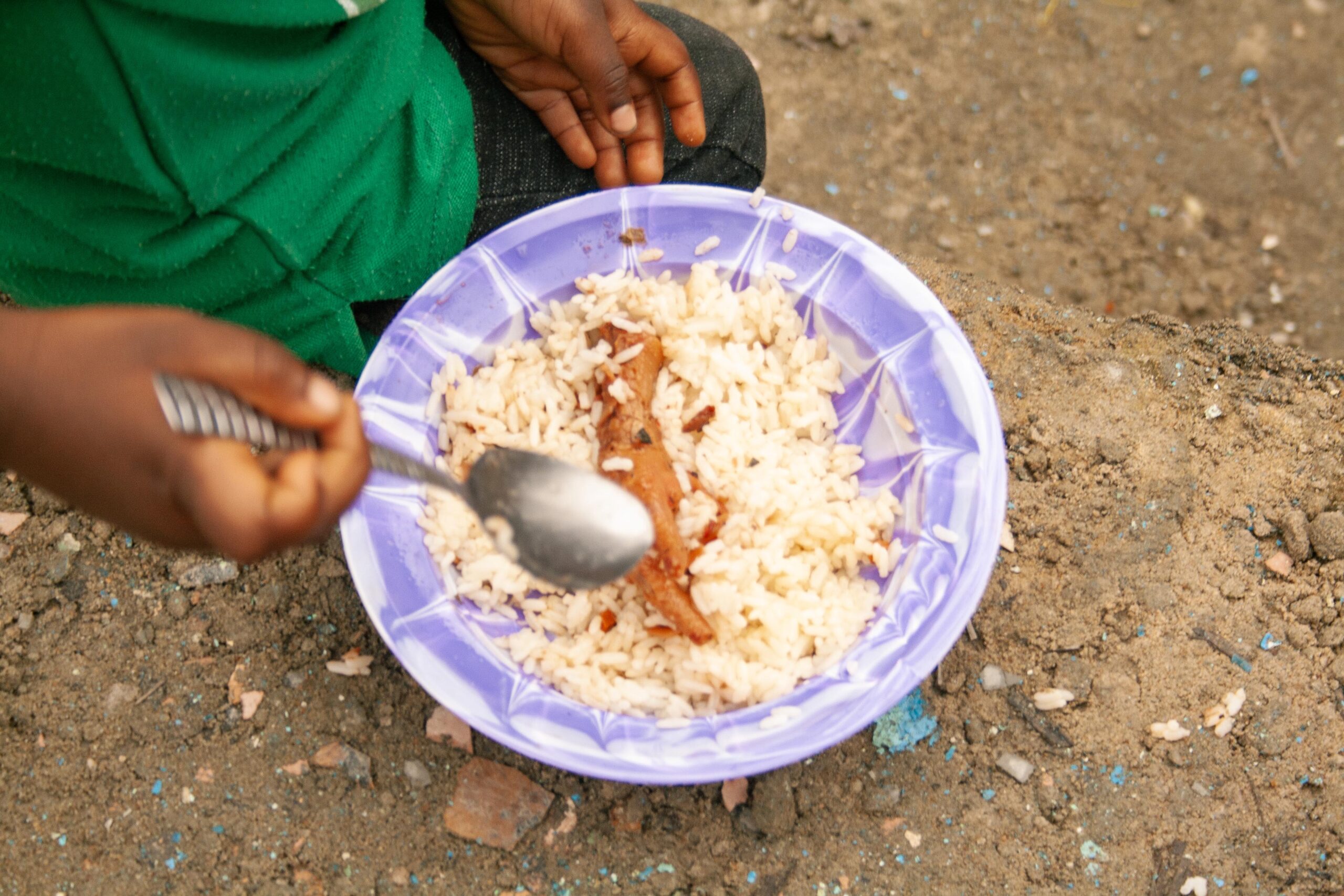 Child holding a spoon while eating rice with sauce from a purple bowl, sitting on the ground. The image highlights the simplicity of a meal in a natural outdoor setting.