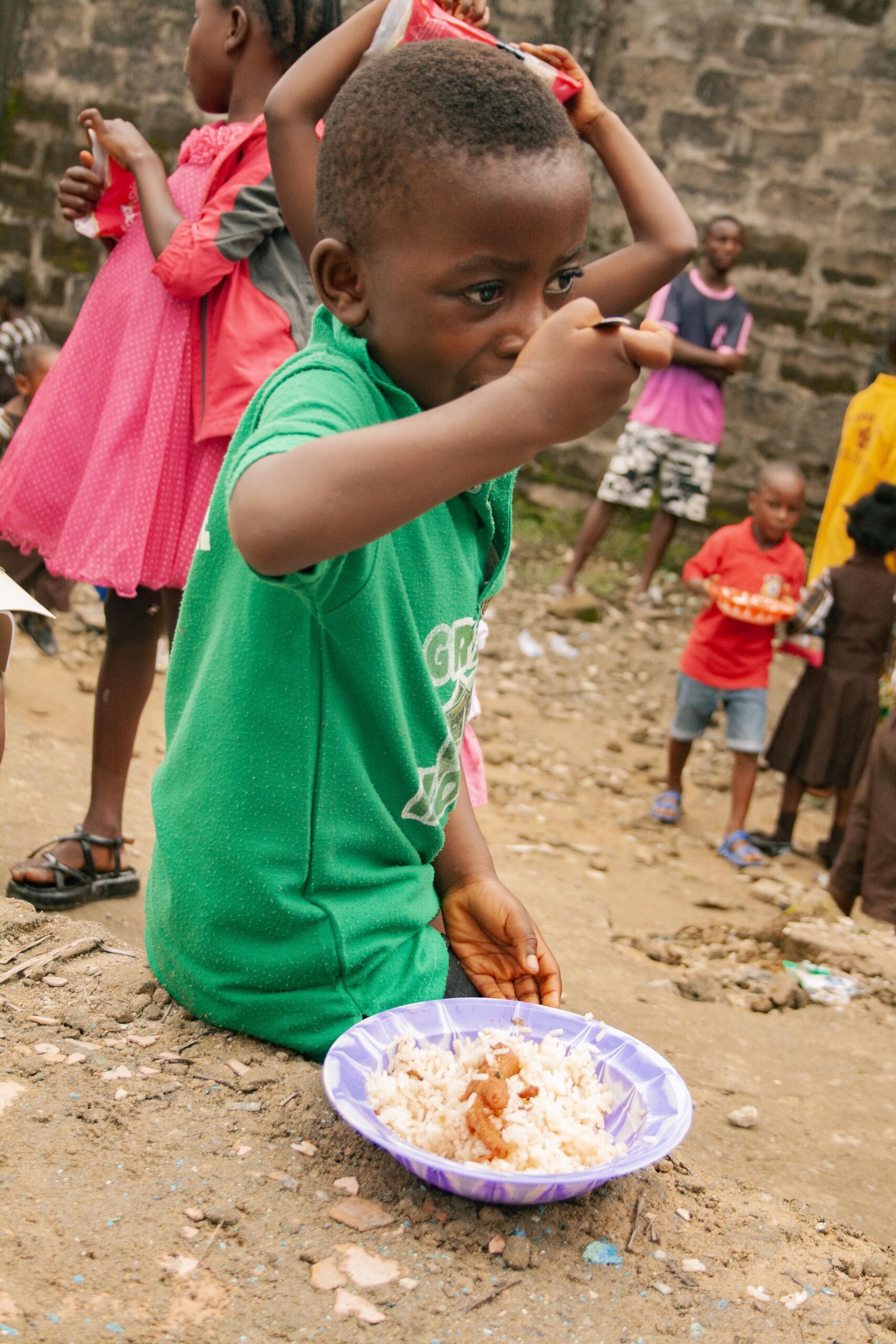 Young child in a green shirt sitting on the ground, eating rice with beans from a purple plate, surrounded by other children in a community setting. The environment appears informal and rugged, highlighting everyday life in a low-income area.