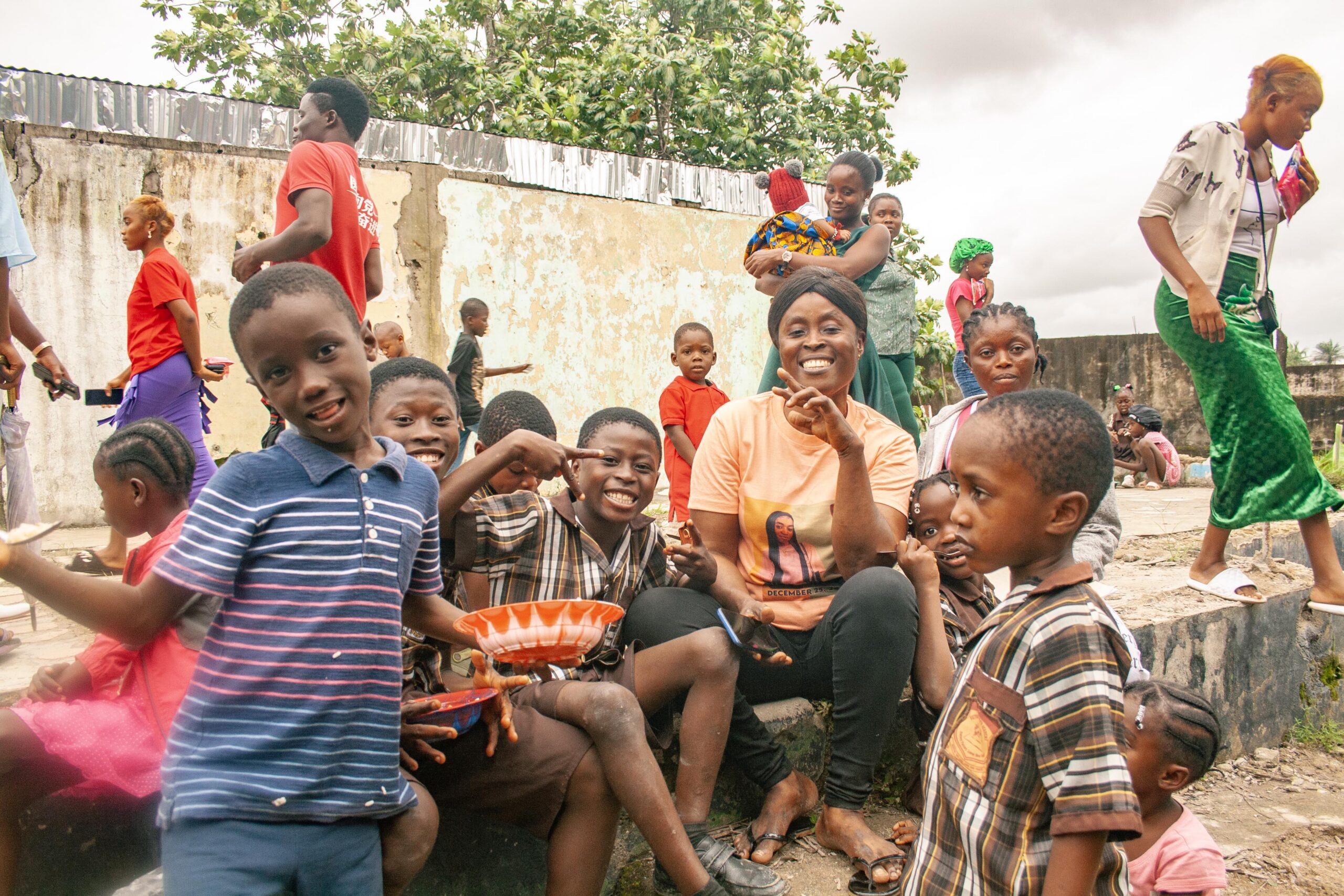 A joyful group of children and adults gather outdoors, smiling and interacting in a community setting. The scene captures a diverse mix of individuals, including a woman in an orange shirt and several children holding bowls, amidst a backdrop of a weathered wall and greenery. The atmosphere conveys a sense of community and togetherness.