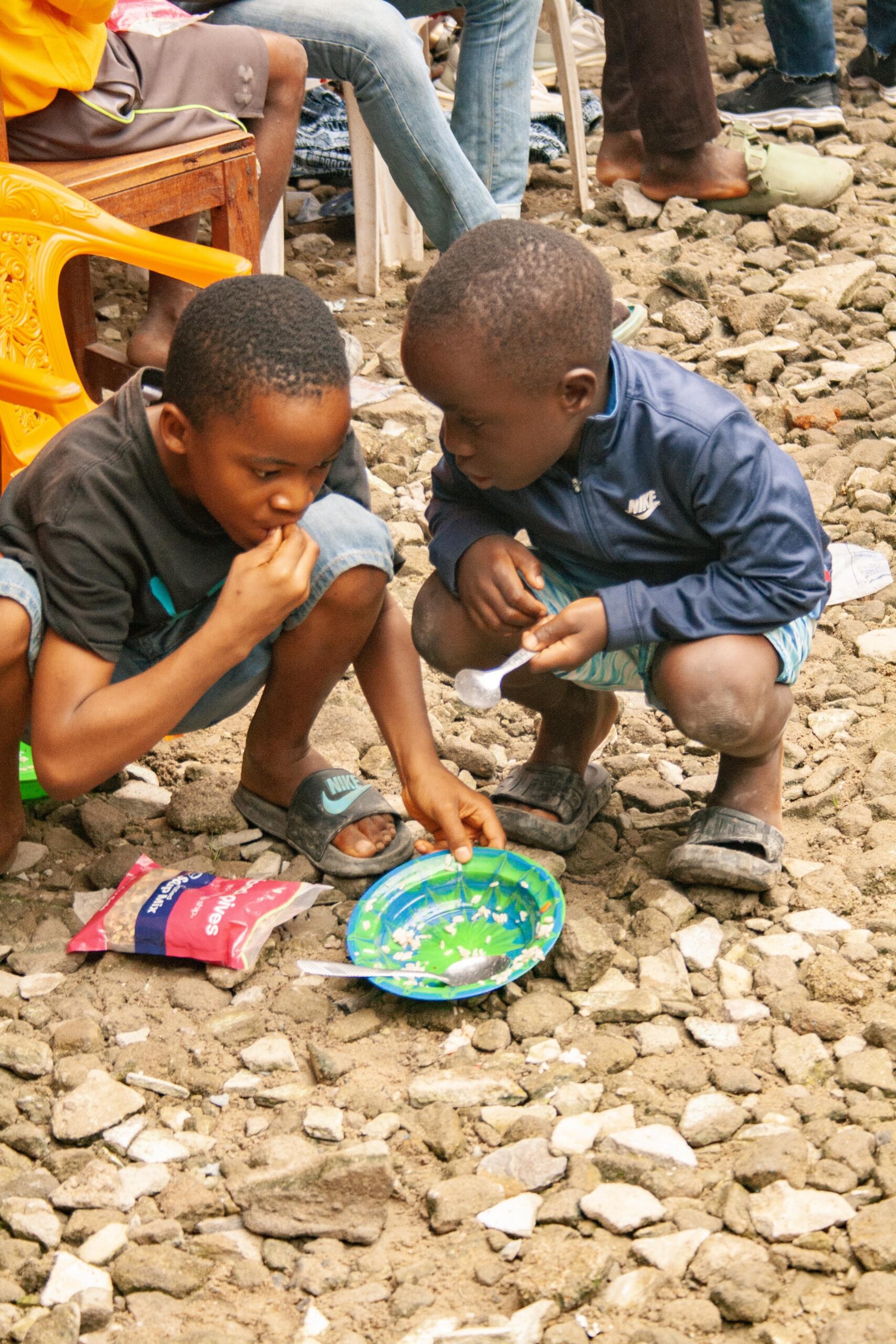 Two young boys share a meal on a colorful plate while sitting on a rocky ground, surrounded by a casual gathering of people. One boy is focused on eating, while the other is holding a spoon, highlighting their friendship and sharing moment.