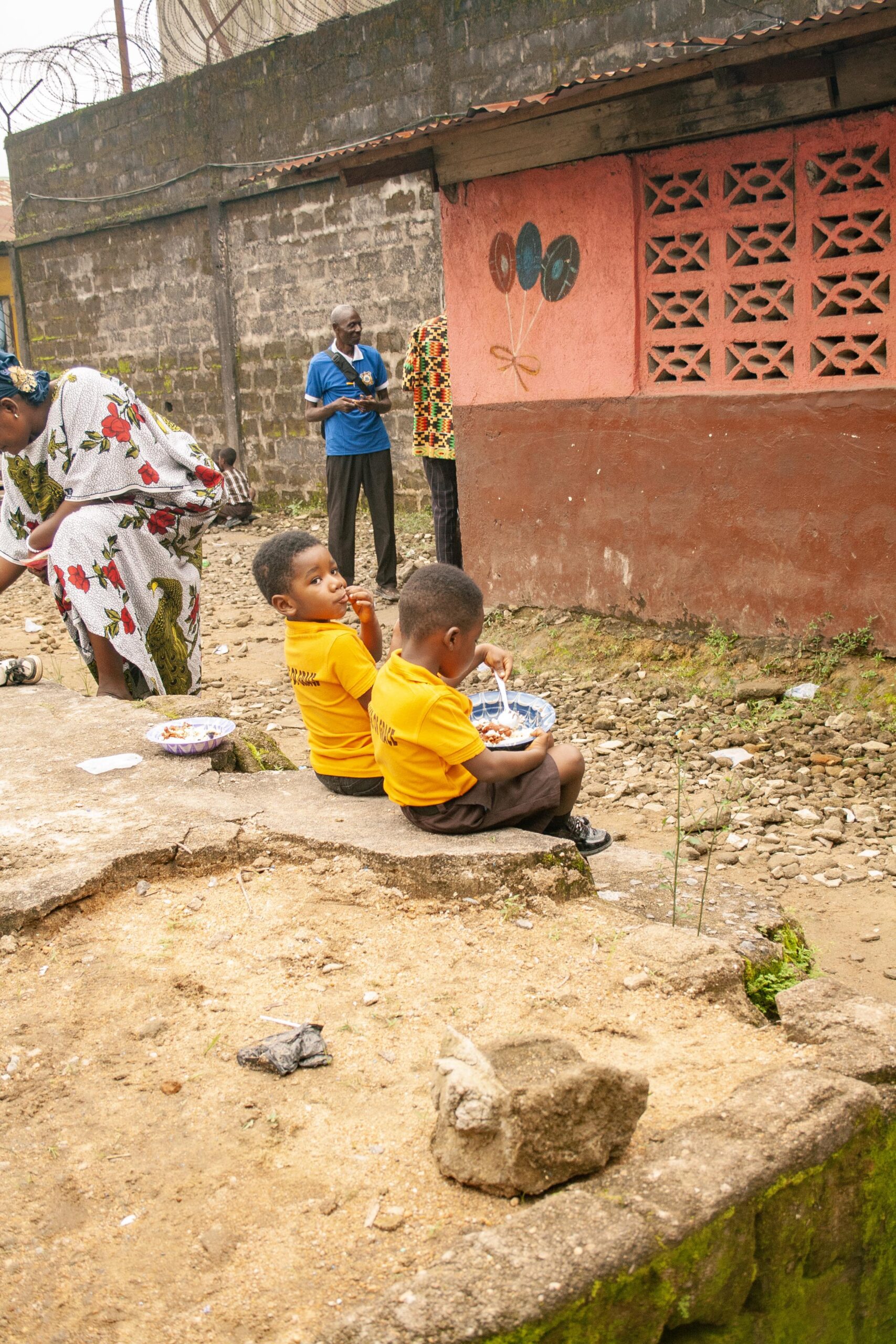 Two young boys in yellow shirts sit on a stone ledge, enjoying a meal. In the background, a woman in a floral dress tends to a task, while adults engage nearby against a backdrop of a colorful wall. The scene captures community life in an urban setting.