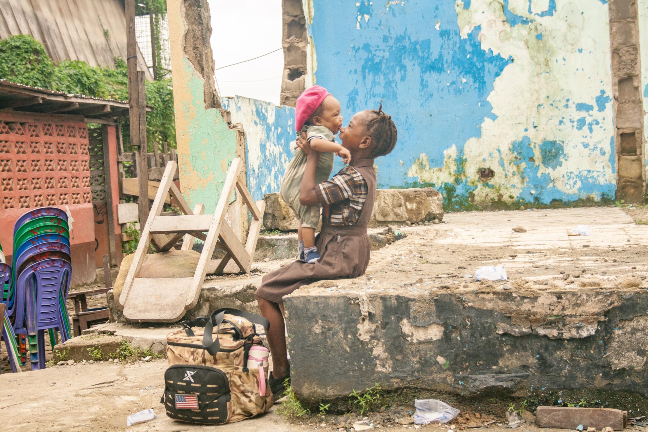 A young girl joyfully holds a toddler in a vibrant urban setting, with colorful walls and scattered debris in the background. The scene captures the bond of love and playfulness amidst a rundown environment, highlighting the resilience of childhood in challenging conditions.