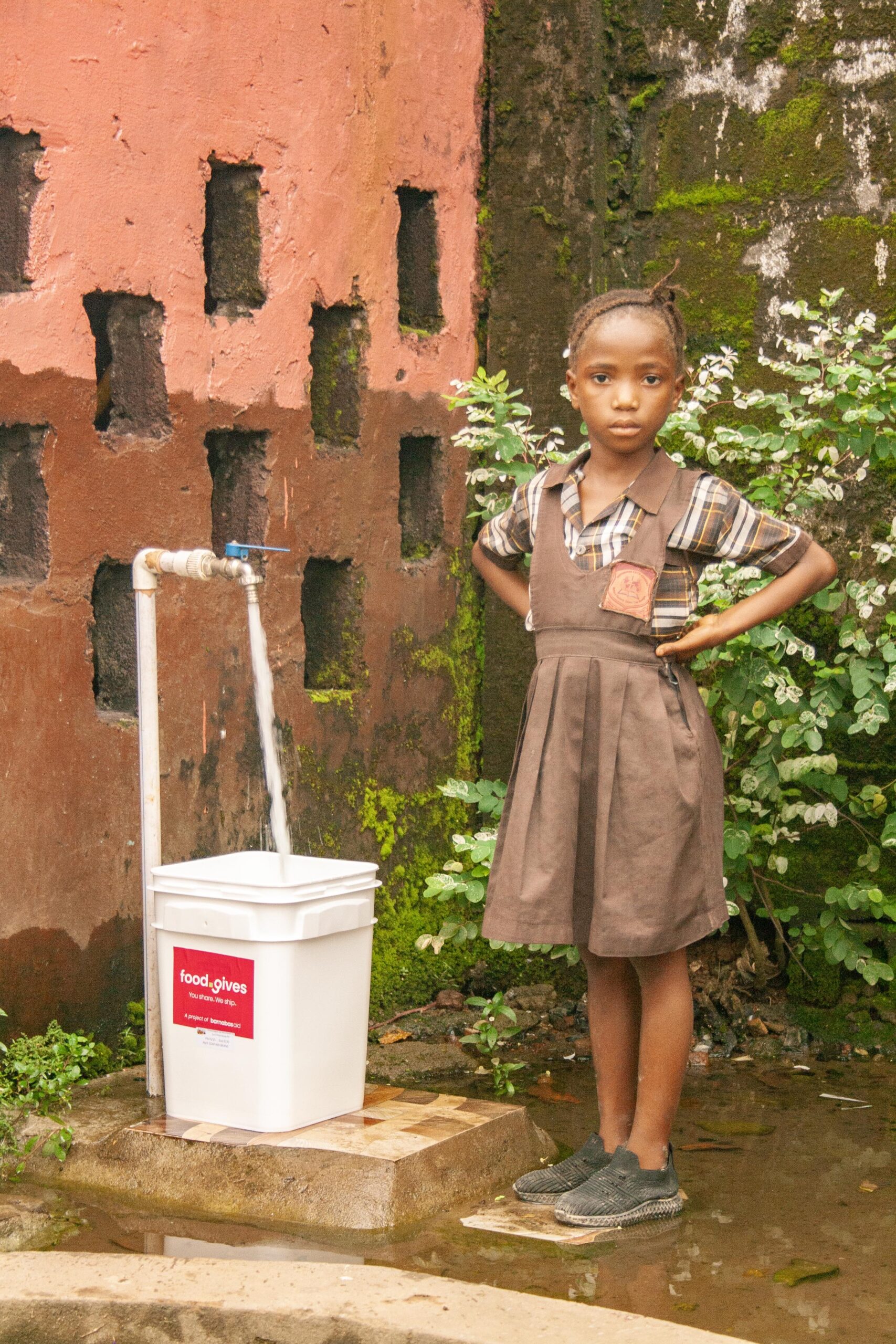 Young girl in a brown dress stands beside a water tap filling a white container, set against a colorful wall with greenery. The image highlights access to clean water in a community setting.