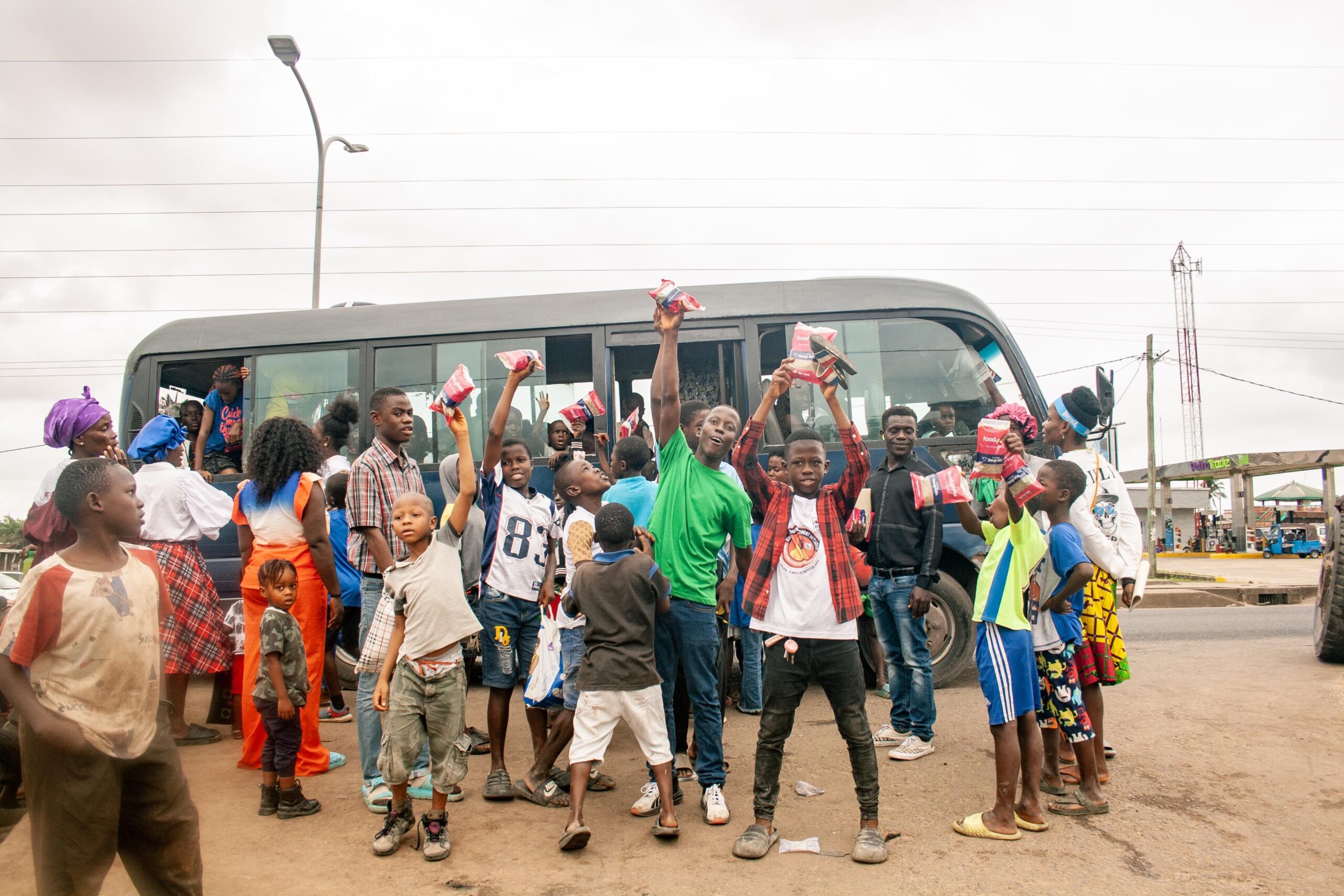 A lively scene of children and adults gathered around a bus, celebrating with joy as they hold up colorful packets. The group, predominantly children, showcases a sense of community and excitement, highlighting a festive atmosphere in an urban setting.