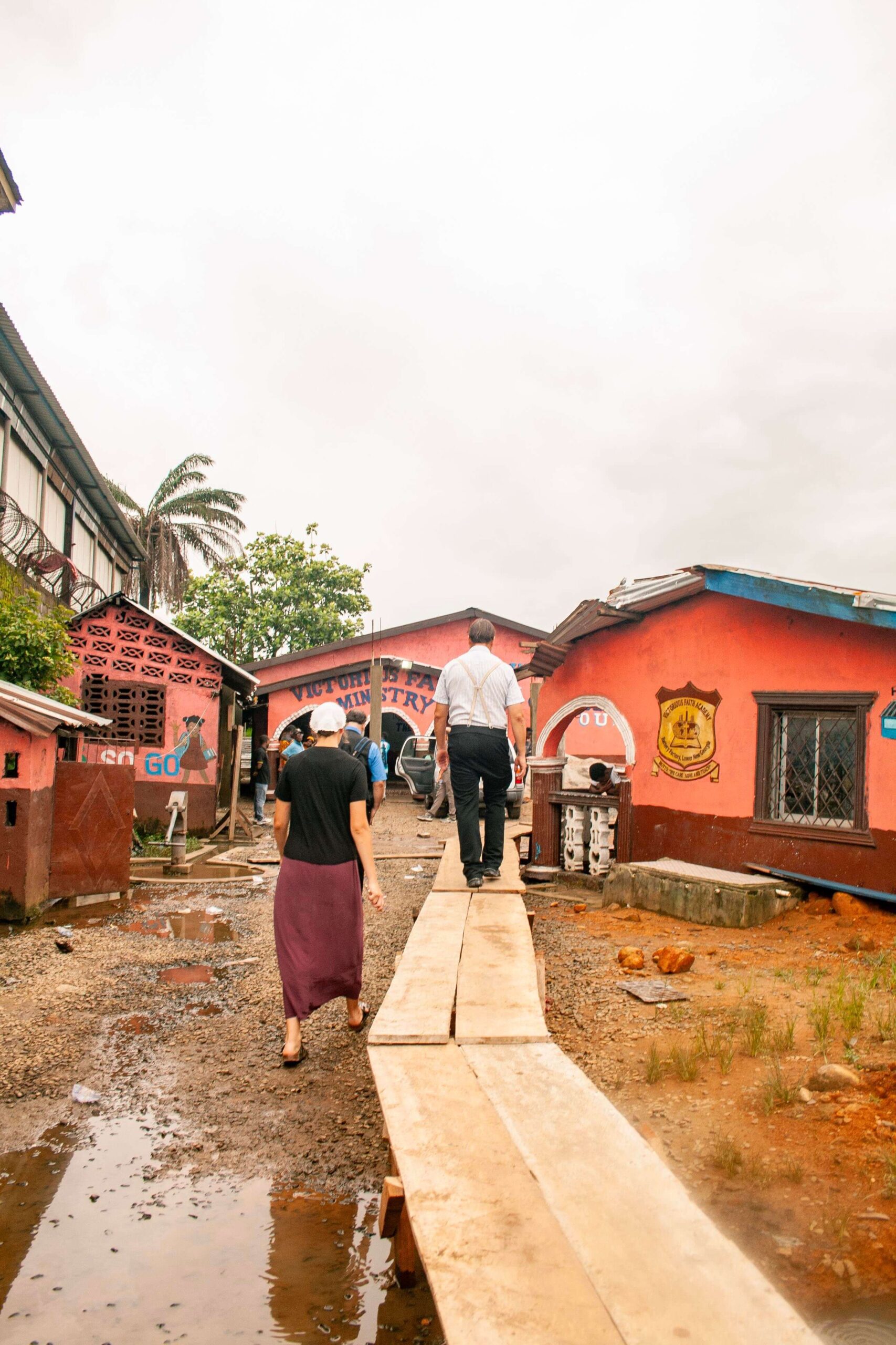 People walking along a wooden pathway in a vibrant community with colorful buildings, palm trees, and a cloudy sky, showcasing local life and culture.