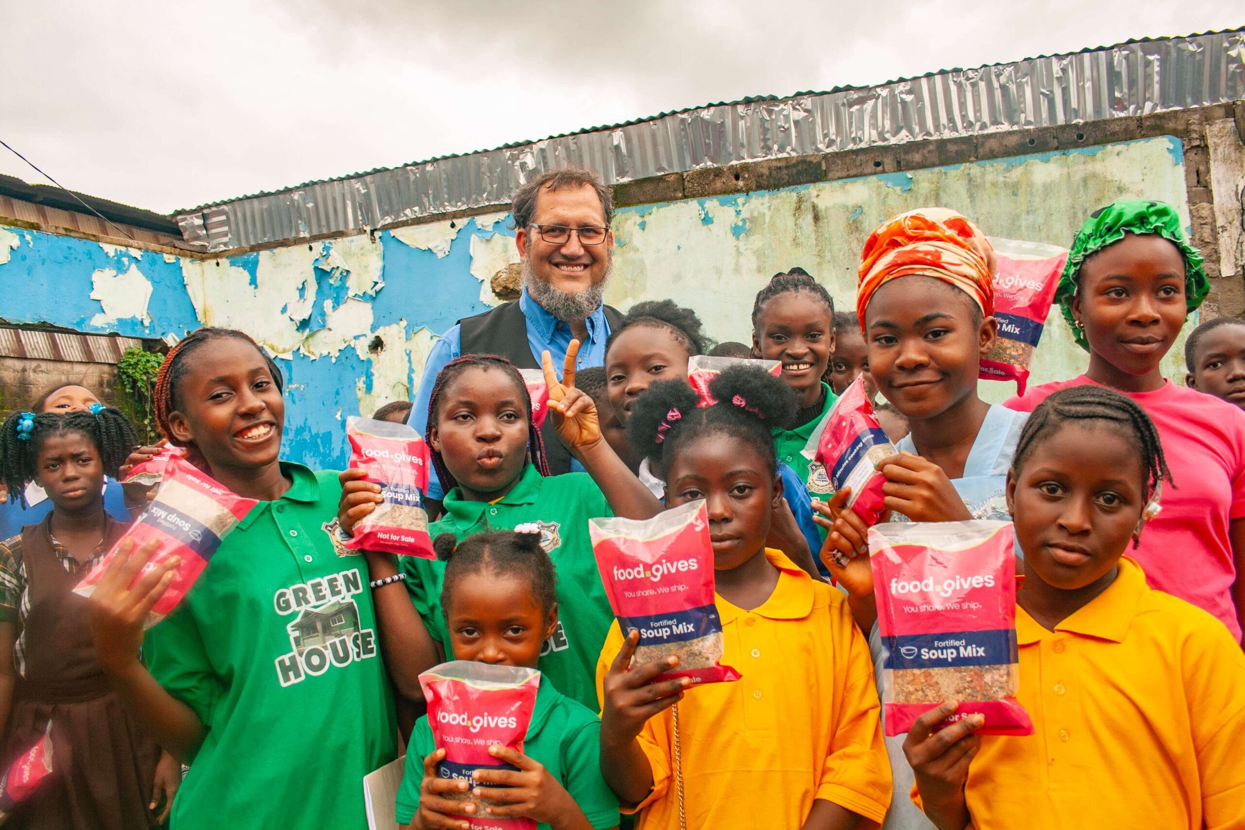 Group of children holding food packages in a community setting, smiling and posing for the camera, with a man in the background. The image highlights a food distribution initiative, showcasing the importance of community support and nutrition.