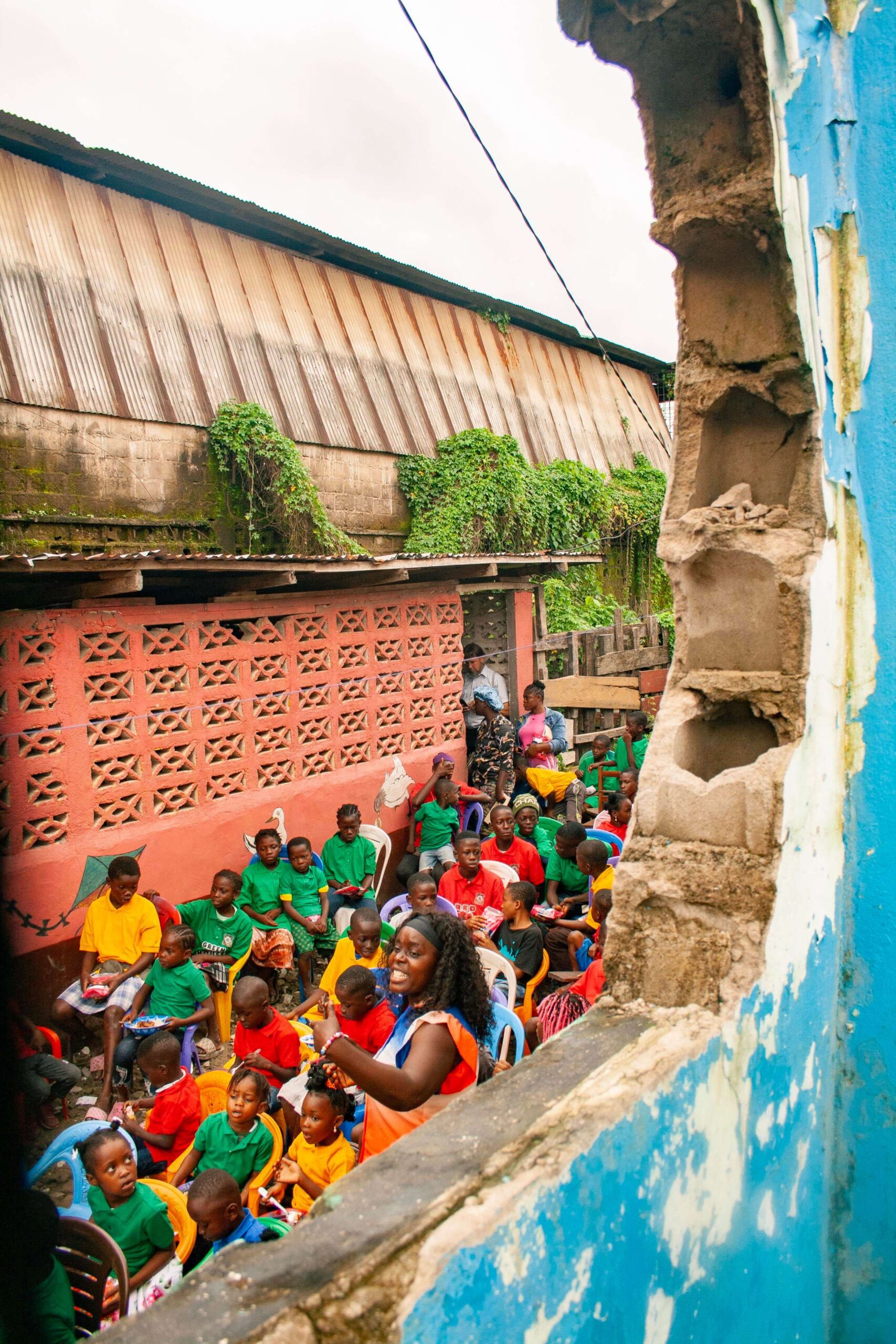 Children in colorful shirts gather in an outdoor classroom setting, engaged with an enthusiastic instructor. The backdrop features a weathered wall covered in greenery, illustrating a community learning environment.