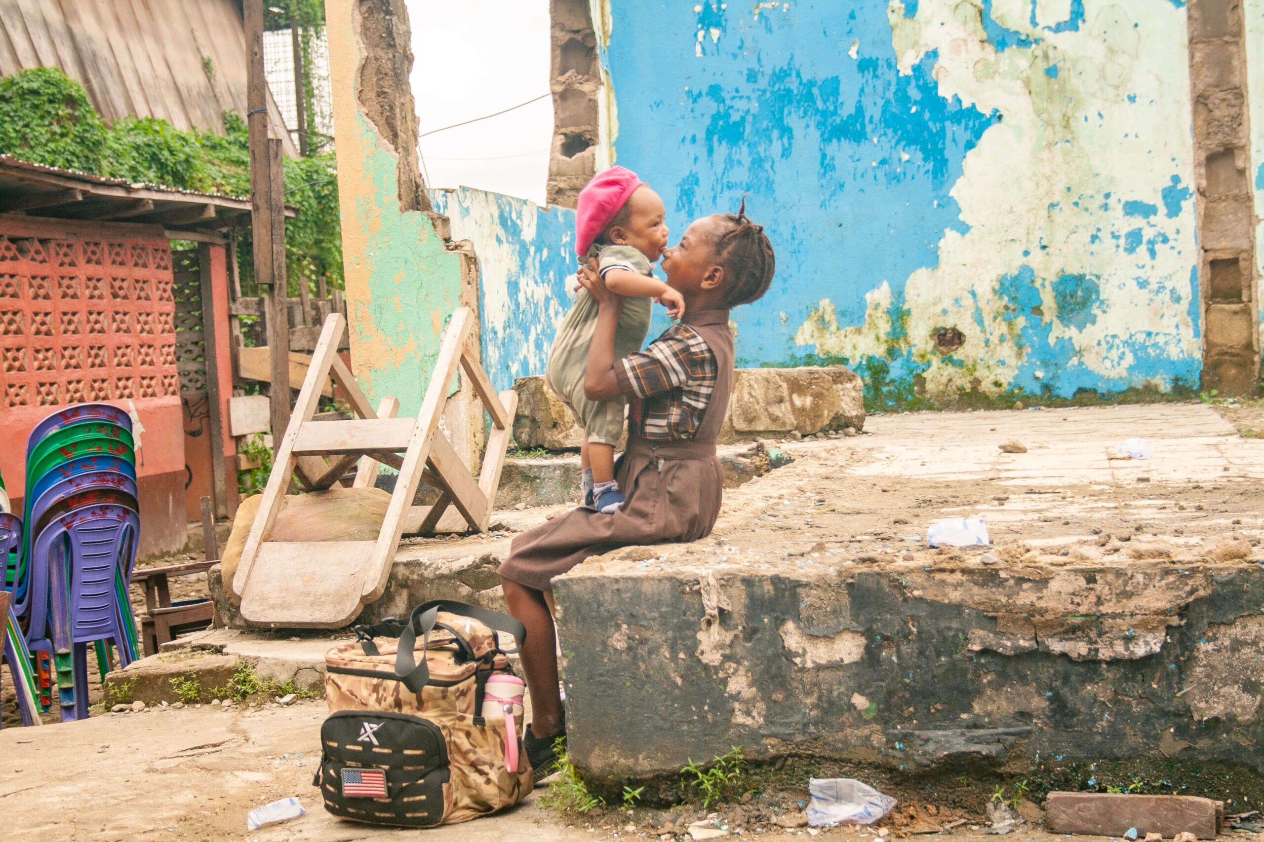 Two children share a joyful moment in an urban setting, with one child being lifted by an older sibling. The background features a weathered wall and scattered debris, highlighting the vibrant yet challenging environment. A backpack is visible on the ground, suggesting a sense of daily life in the area.