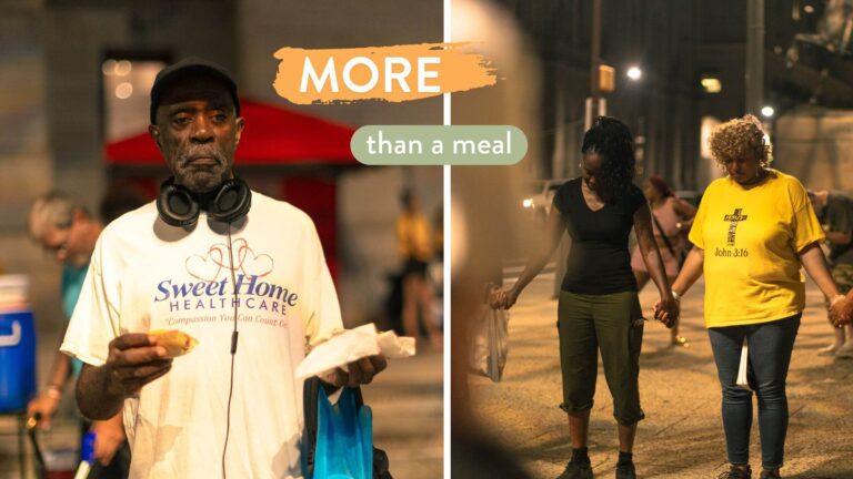 A man holding food stands in the foreground, wearing headphones and a "Sweet Home Healthcare" shirt, on a summer evening in Philadelphia, while two women in the background hold hands and pray together, symbolizing community support and compassion through more than a meal.