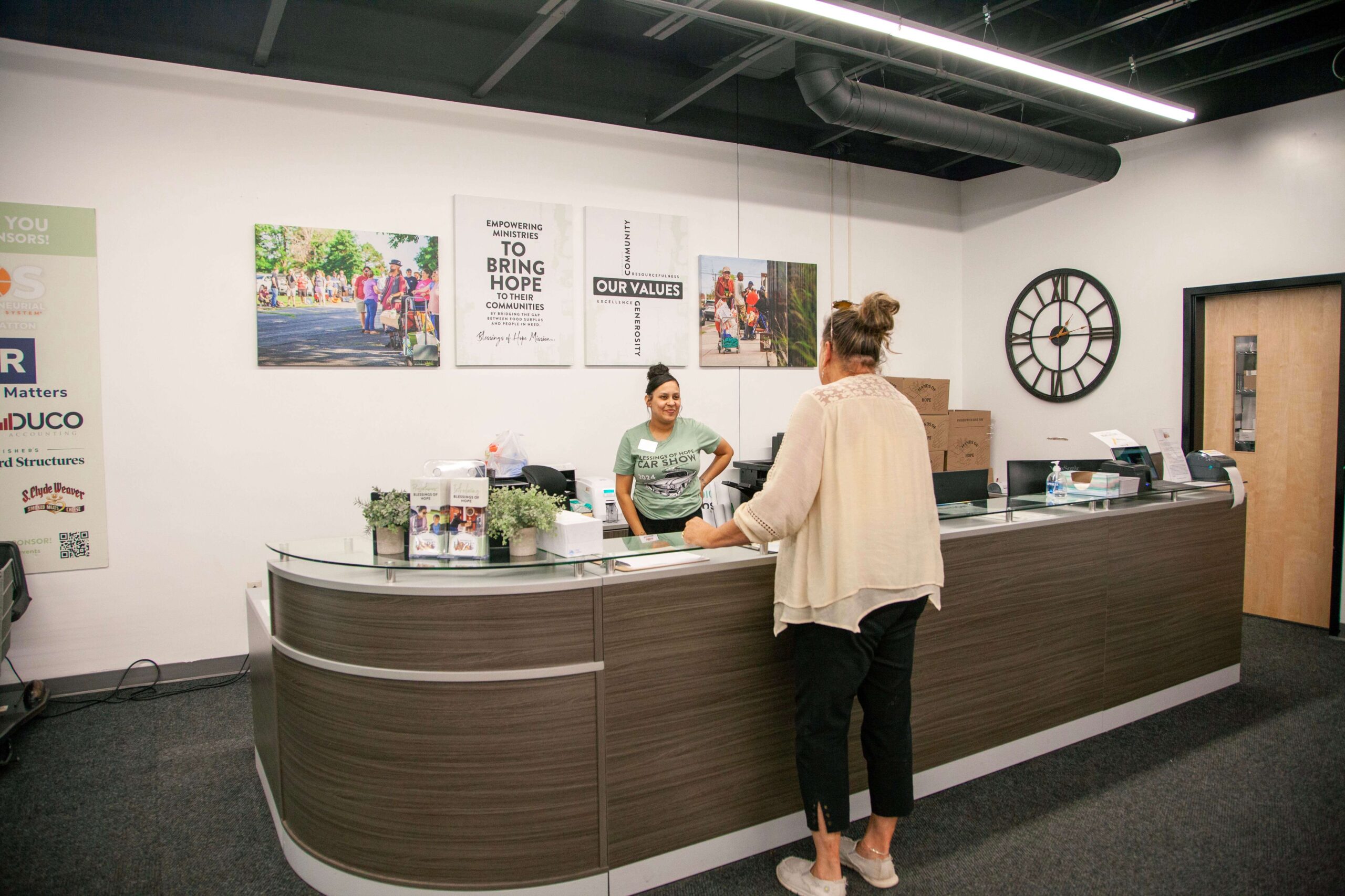 Reception area of a community service organization featuring a welcoming staff member at the front desk, surrounded by informational displays and community-focused artwork on the walls.