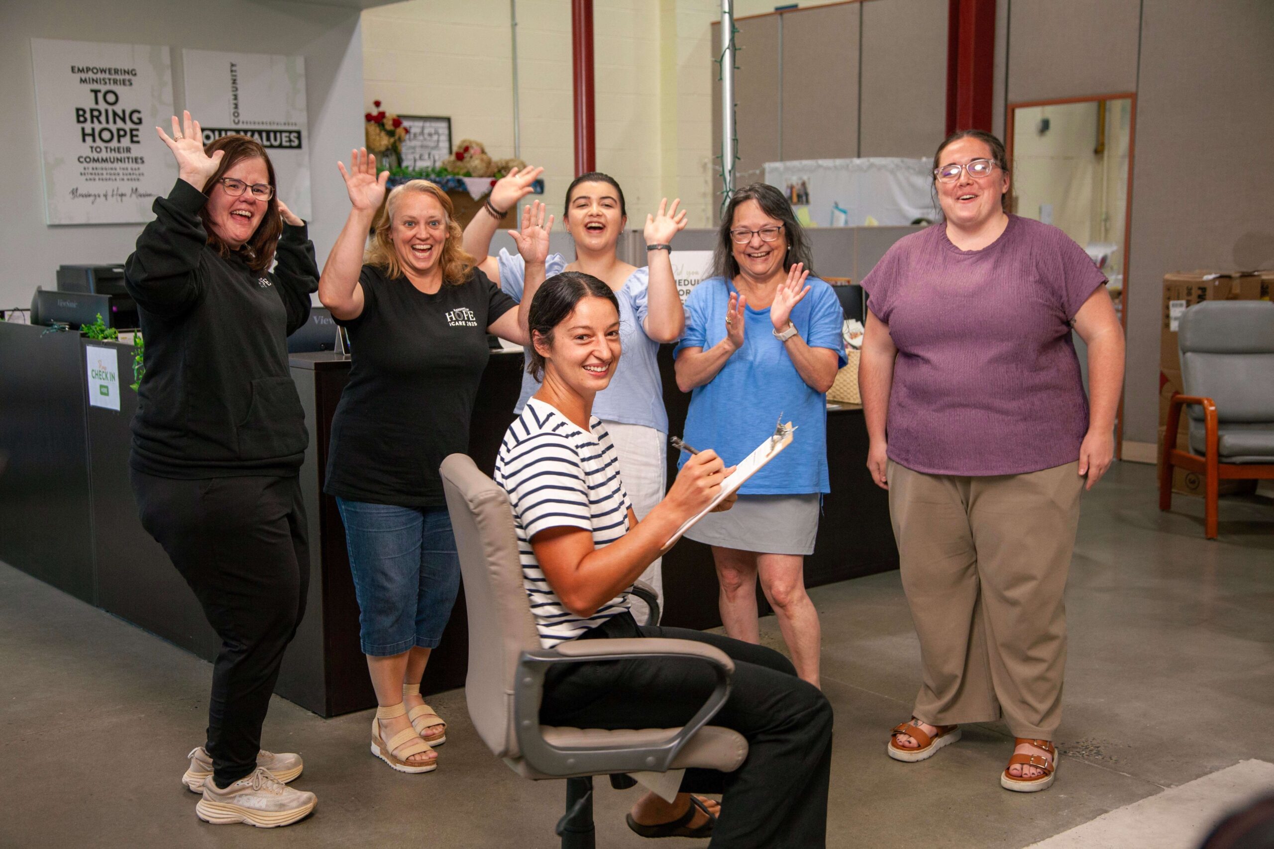 A group of six women enthusiastically celebrate in an office setting, with one woman seated and taking notes. The background features signs promoting community empowerment and hope.