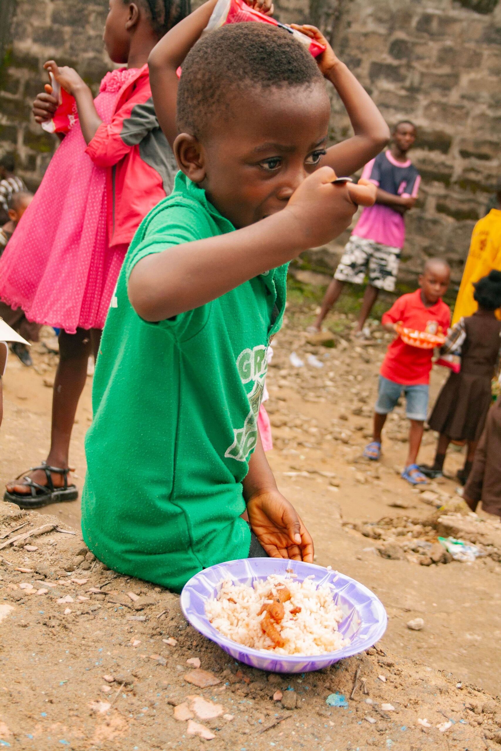 A young child in a green shirt sits on the ground, enjoying a bowl of rice in a vibrant community setting, surrounded by other children playing in the background. The image highlights themes of childhood, community, and food sharing.