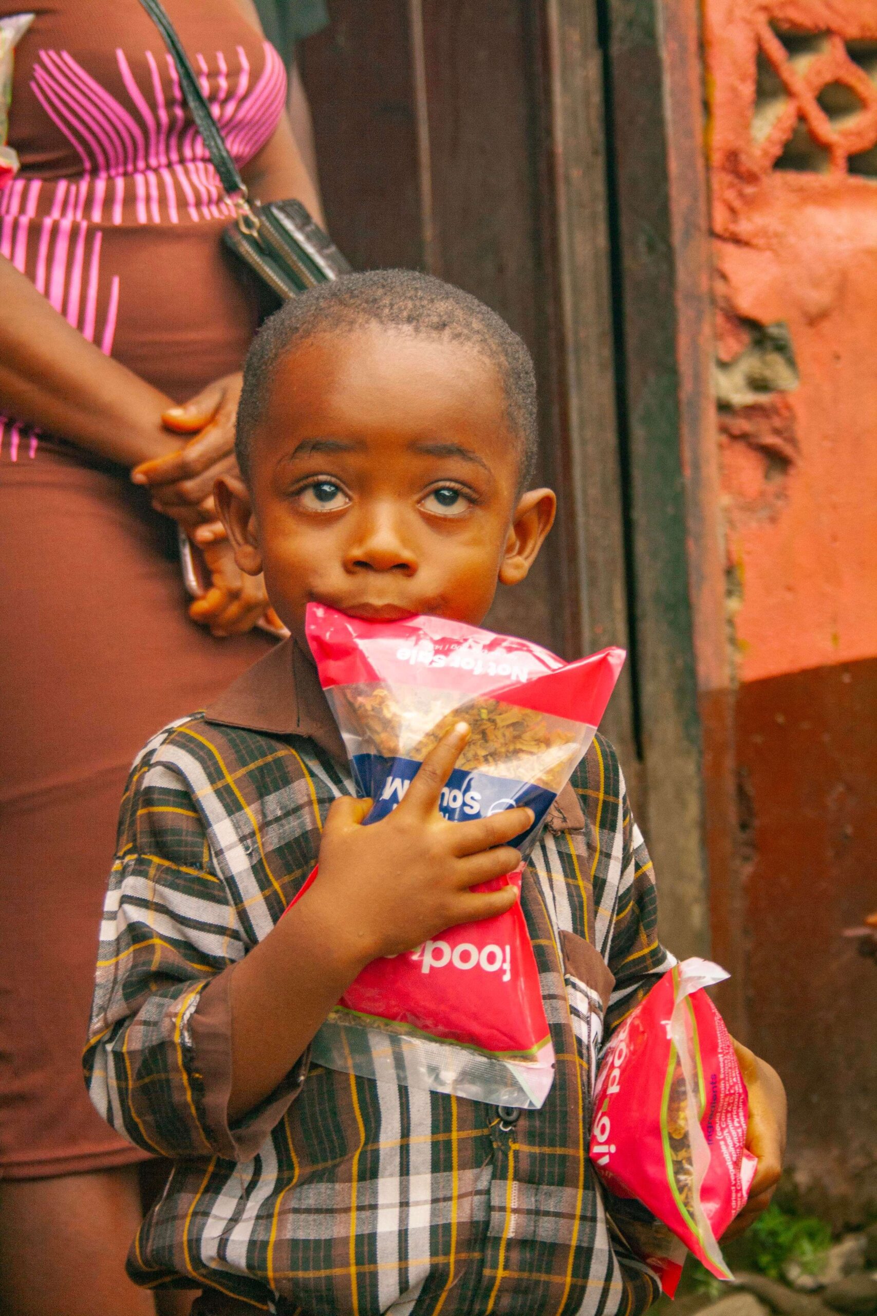Young boy holding packets of snacks in a vibrant street setting, with an adult figure in the background. The child has expressive blue eyes and is dressed in a plaid shirt, capturing a moment of joy and curiosity.