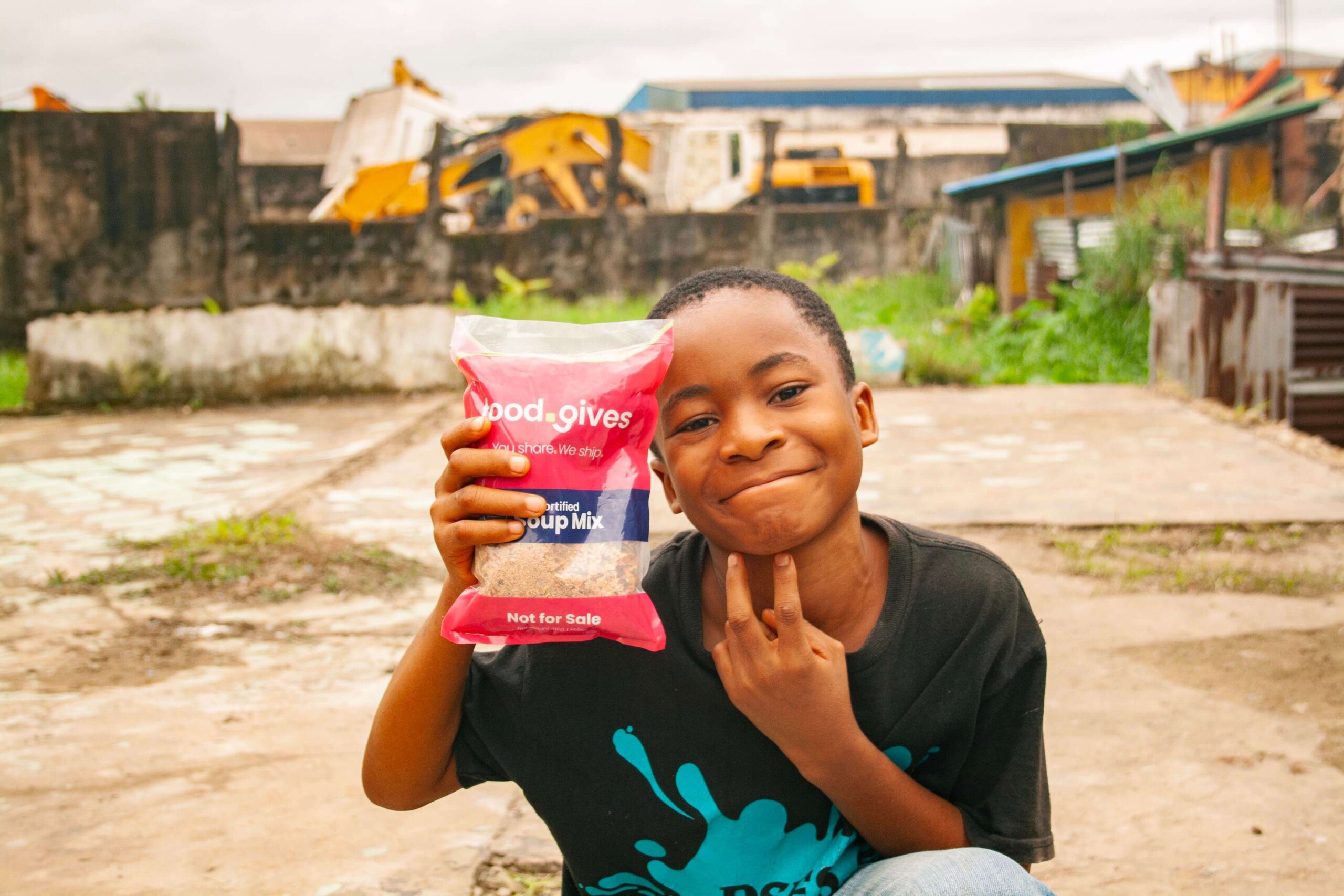 Smiling boy holding a packet of food soup mix labeled "food.gives," promoting community sharing and support in an outdoor setting with construction equipment in the background.