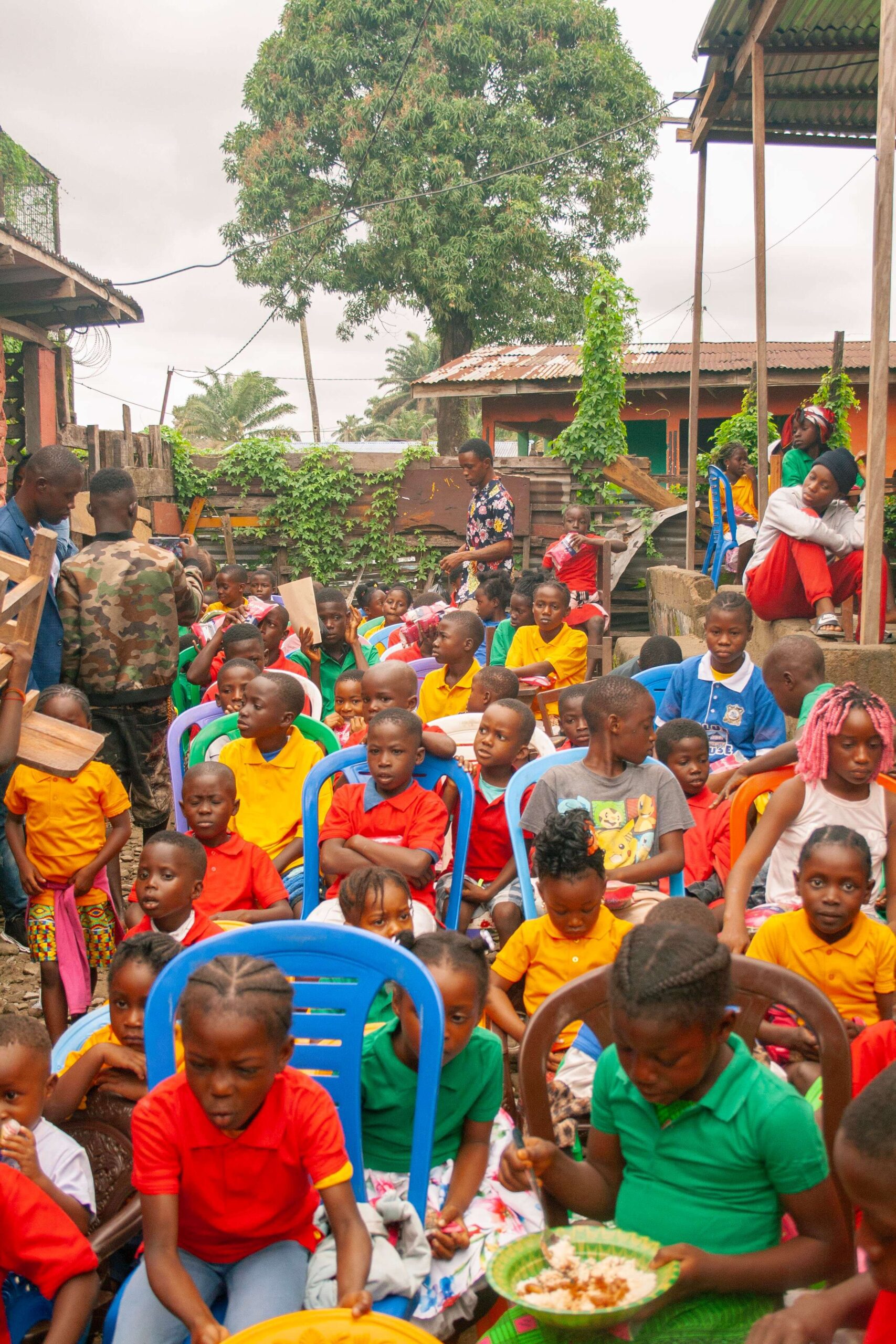 A vibrant outdoor gathering of children seated on plastic chairs, wearing colorful shirts, enjoying a meal in a community setting. The scene features a diverse group of kids with various hairstyles, engaged in a joyful event, surrounded by greenery and informal structures.