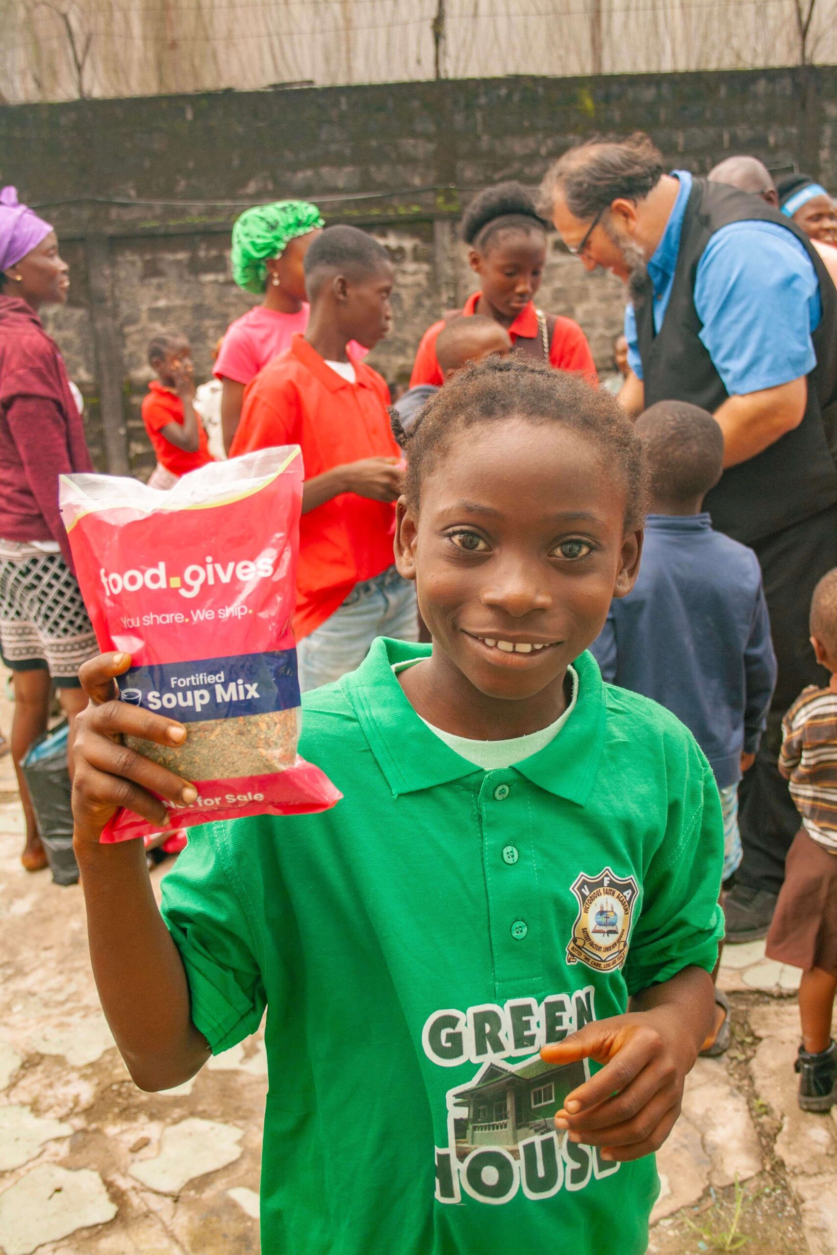 Smiling child in a green shirt holding a packet of fortified soup mix, surrounded by a community event with people in colorful clothing in the background, promoting food assistance and support.
