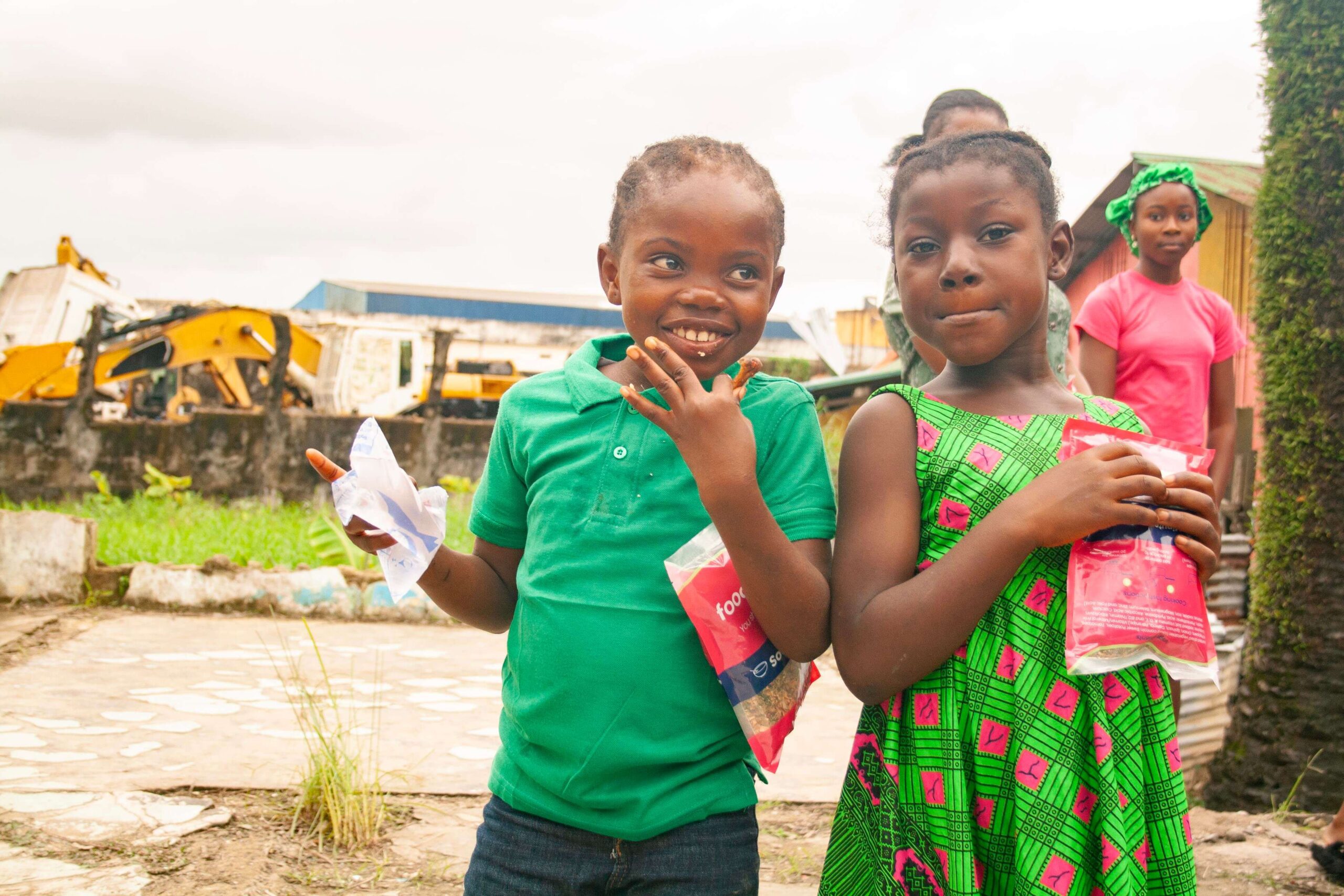 Two smiling children in colorful clothing hold snack packages, with a backdrop of greenery and construction vehicles. The scene captures a moment of joy and community engagement.