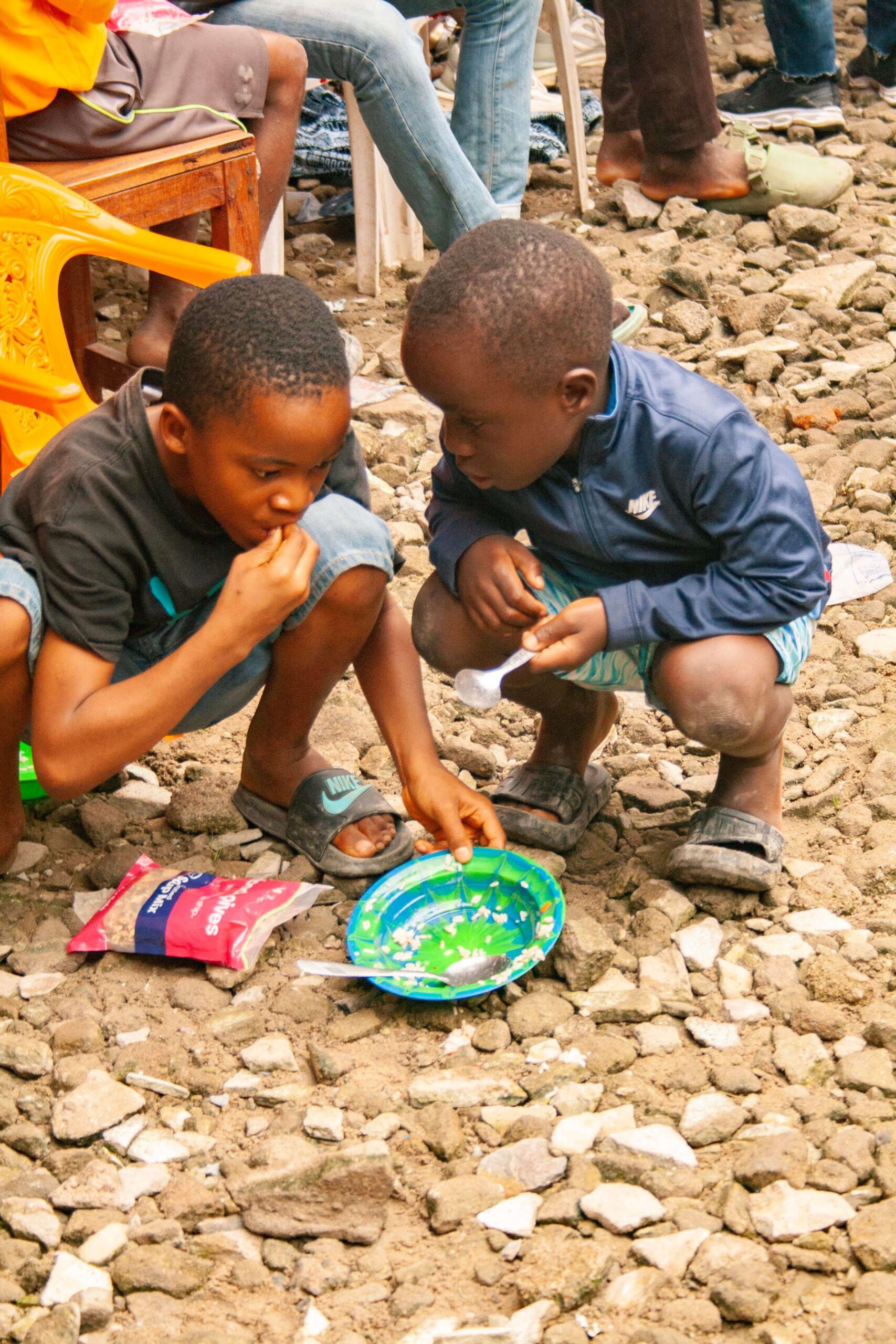 Two young boys sharing food from a colorful plate while sitting on rocky ground, surrounded by a casual outdoor setting. One boy is intently eating, while the other looks on, both dressed in casual clothing.