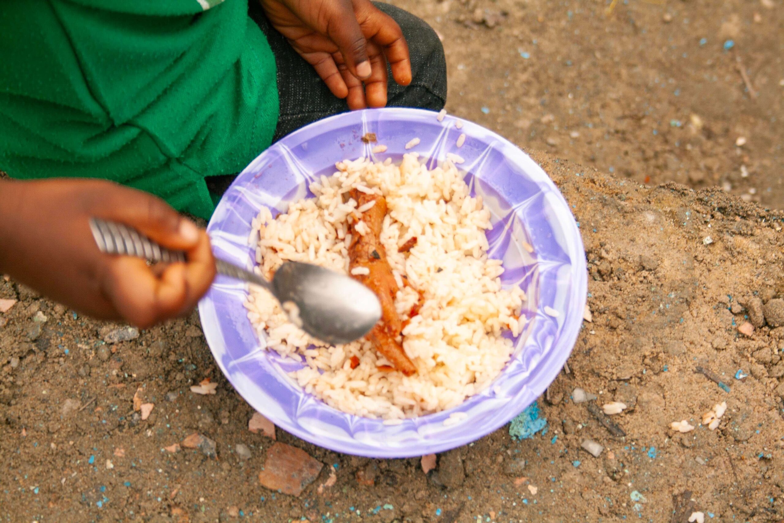Child's hand holding a spoon, mixing rice and sauce in a purple bowl, sitting on the ground. The image highlights a simple meal, emphasizing the importance of nutrition and food accessibility.
