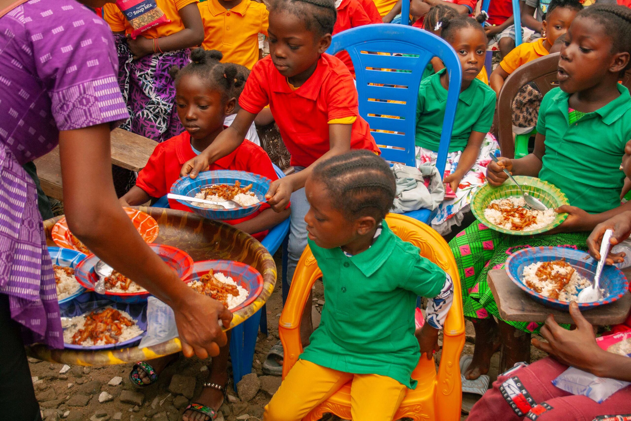 Children seated at a community gathering eagerly receiving plates of rice and stew from a caregiver. The scene showcases a vibrant atmosphere with kids in colorful shirts, highlighting a communal meal in a cheerful outdoor setting.