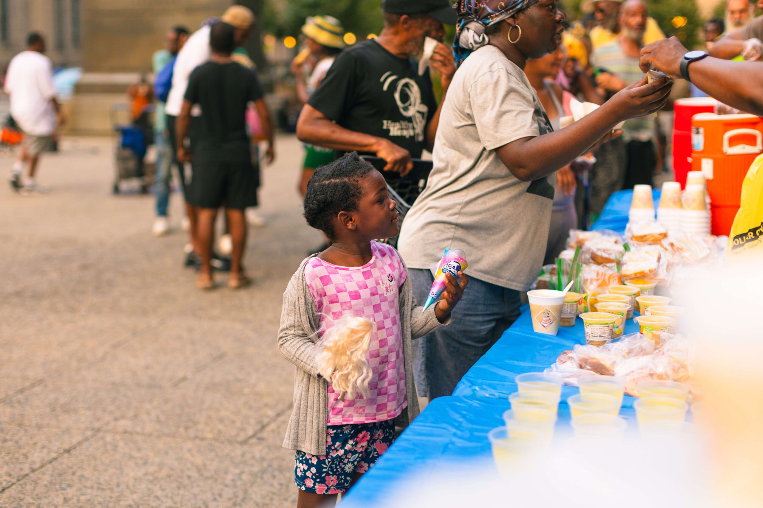 Young girl holding an ice cream cone and a bag of snacks stands beside a table filled with food and drinks at a community event, surrounded by people enjoying the festivities.
