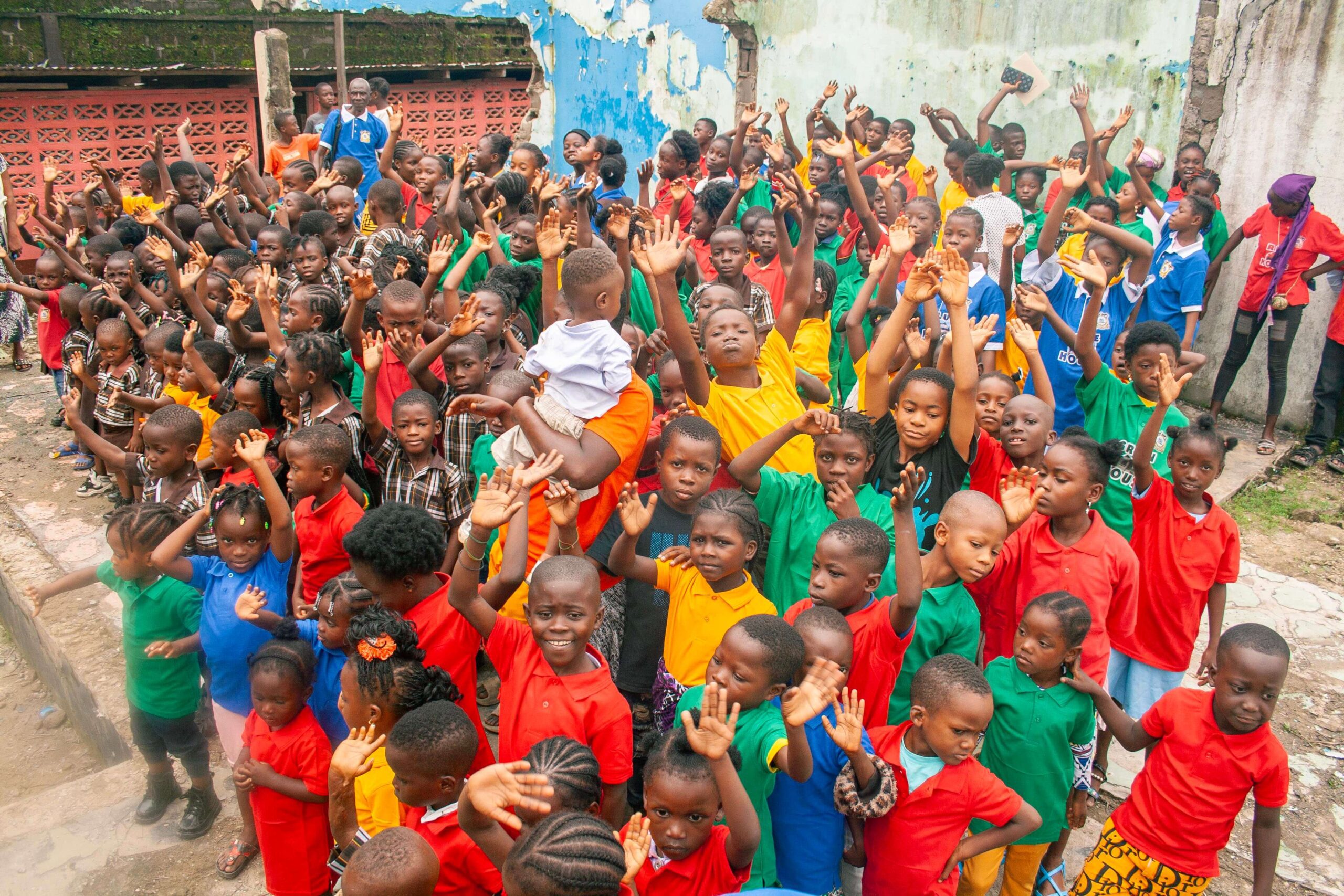 A vibrant group of children in colorful shirts gathered outdoors, raising their hands in celebration or greeting. The scene showcases a diverse crowd of kids, highlighting community engagement and joy in a lively environment.