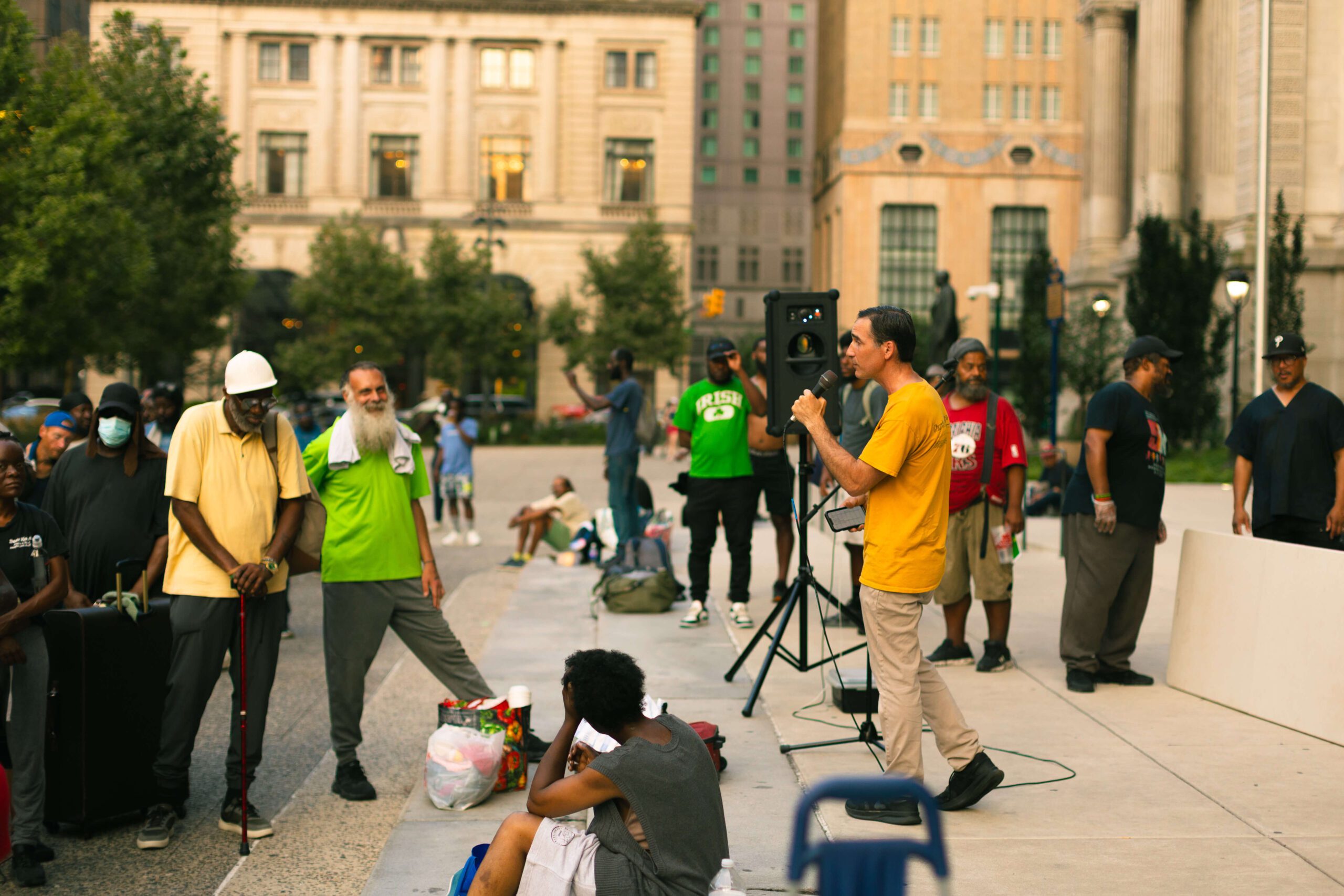 A community gathering in an urban park where a speaker addresses a diverse audience, including men and women of various ages. Some attendees are seated while others stand in support, highlighting themes of unity and social engagement. The backdrop features historical buildings and greenery, emphasizing the event's public setting.