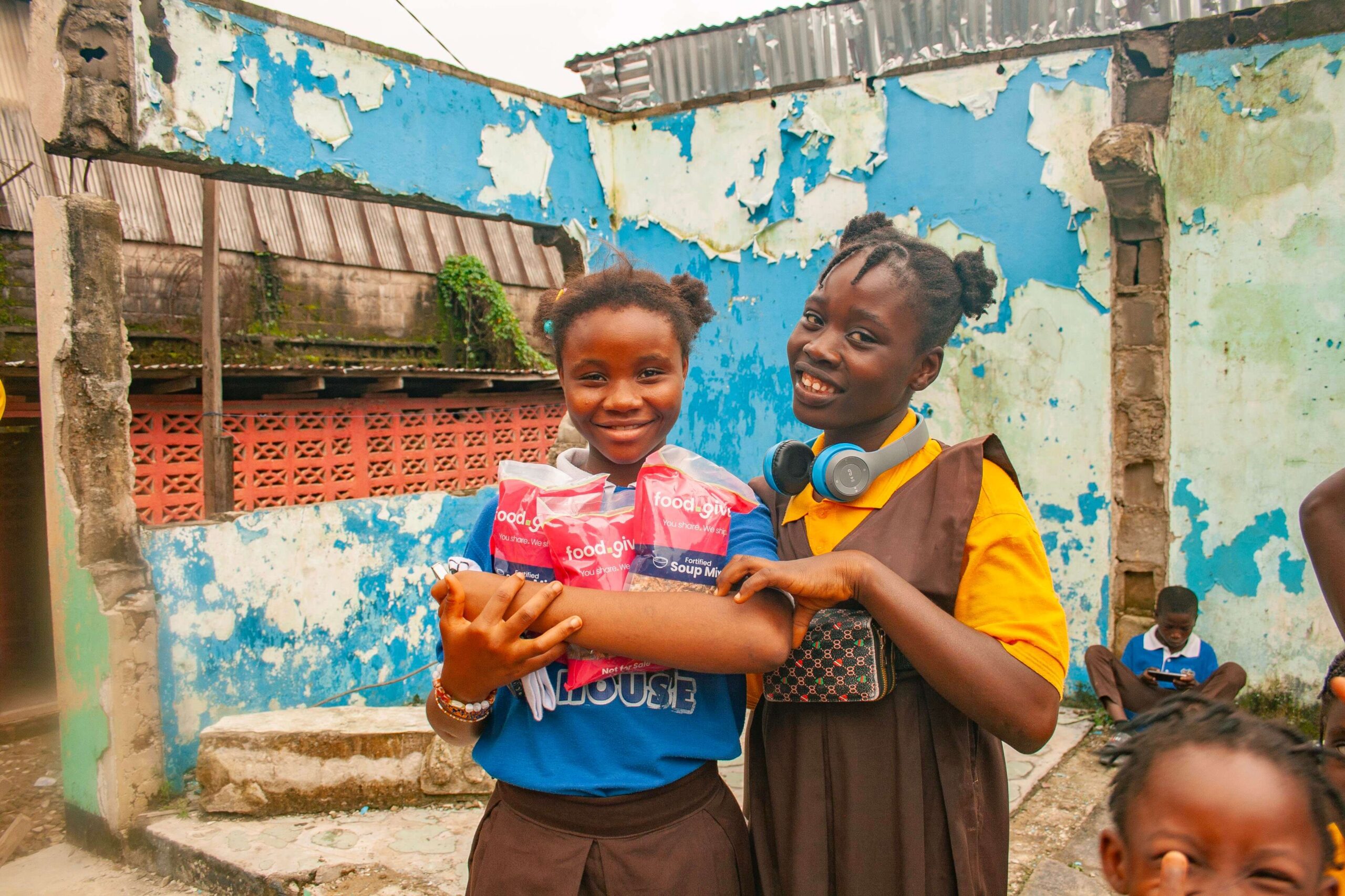 Two smiling girls hold packages of fortified soup mix in a community setting, with a backdrop of peeling blue walls and other children in the background. The image highlights the importance of food aid and community support in underserved areas.