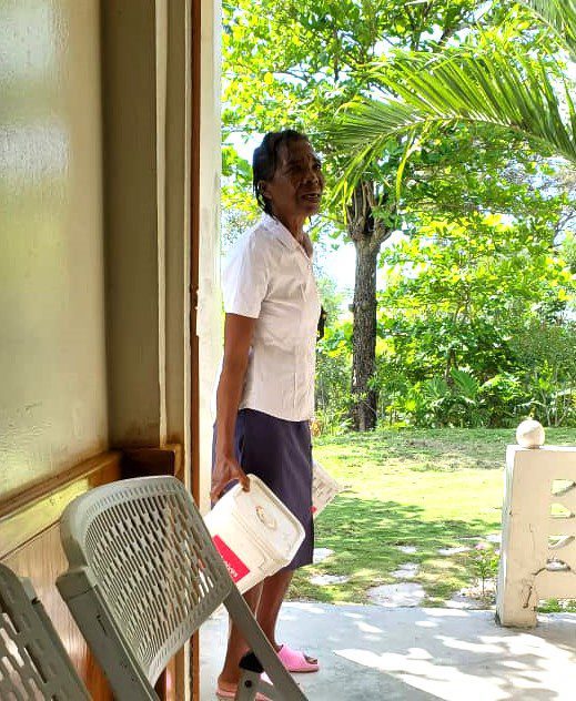 A woman in a white blouse and navy skirt stands at an open door, holding a container in one hand, with a sunny outdoor scene featuring green trees in the background. A plastic chair is visible in the foreground.