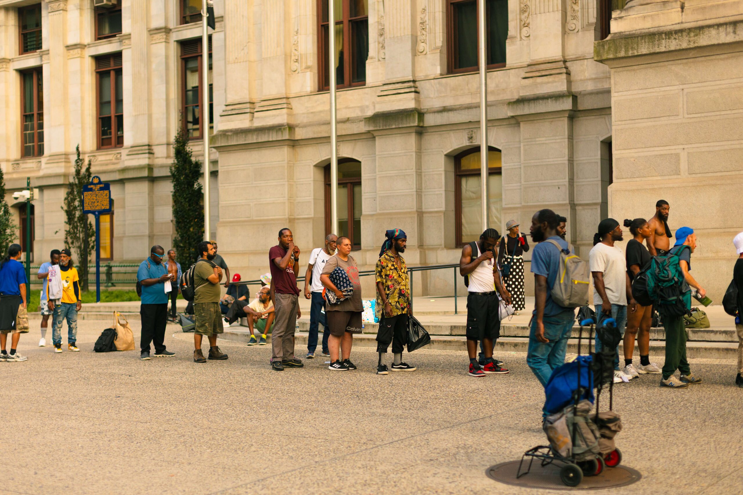 A diverse group of people standing in line outside a historic building, with some individuals engaged in conversation and others holding bags. The scene captures urban life and community interactions in a city setting.