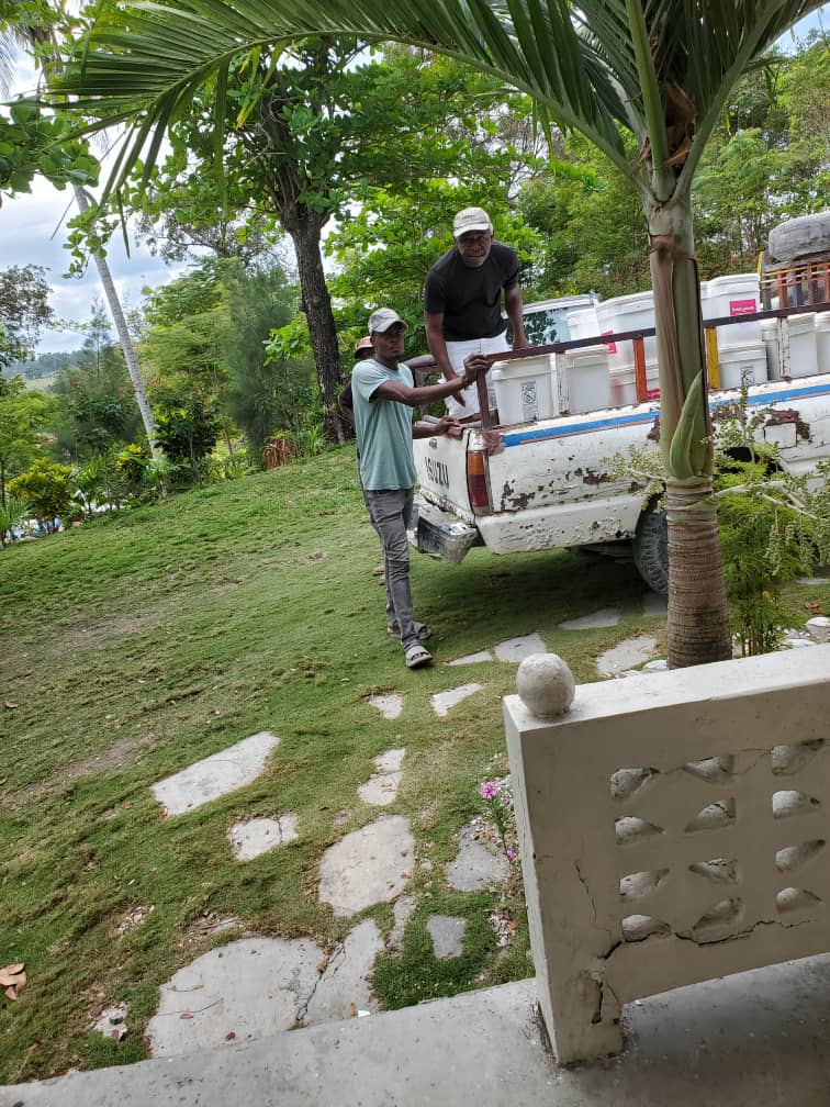 Two men unloading containers from a pickup truck in a green outdoor setting, surrounded by trees and grass. One man is standing in the truck while the other is on the ground, assisting with the unloading process.