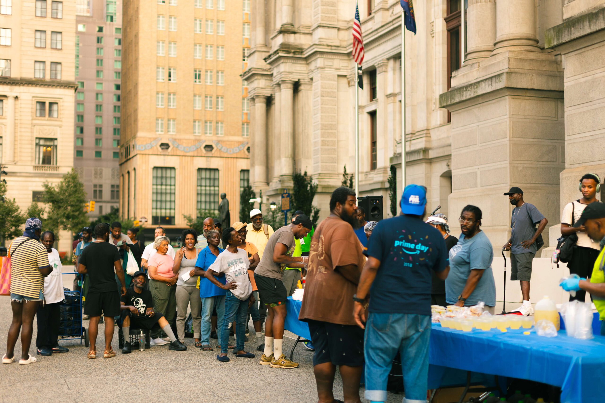 Community members gather at an outdoor event, engaging in conversation and enjoying refreshments at a table filled with food and drinks, set against a backdrop of city buildings and flags.