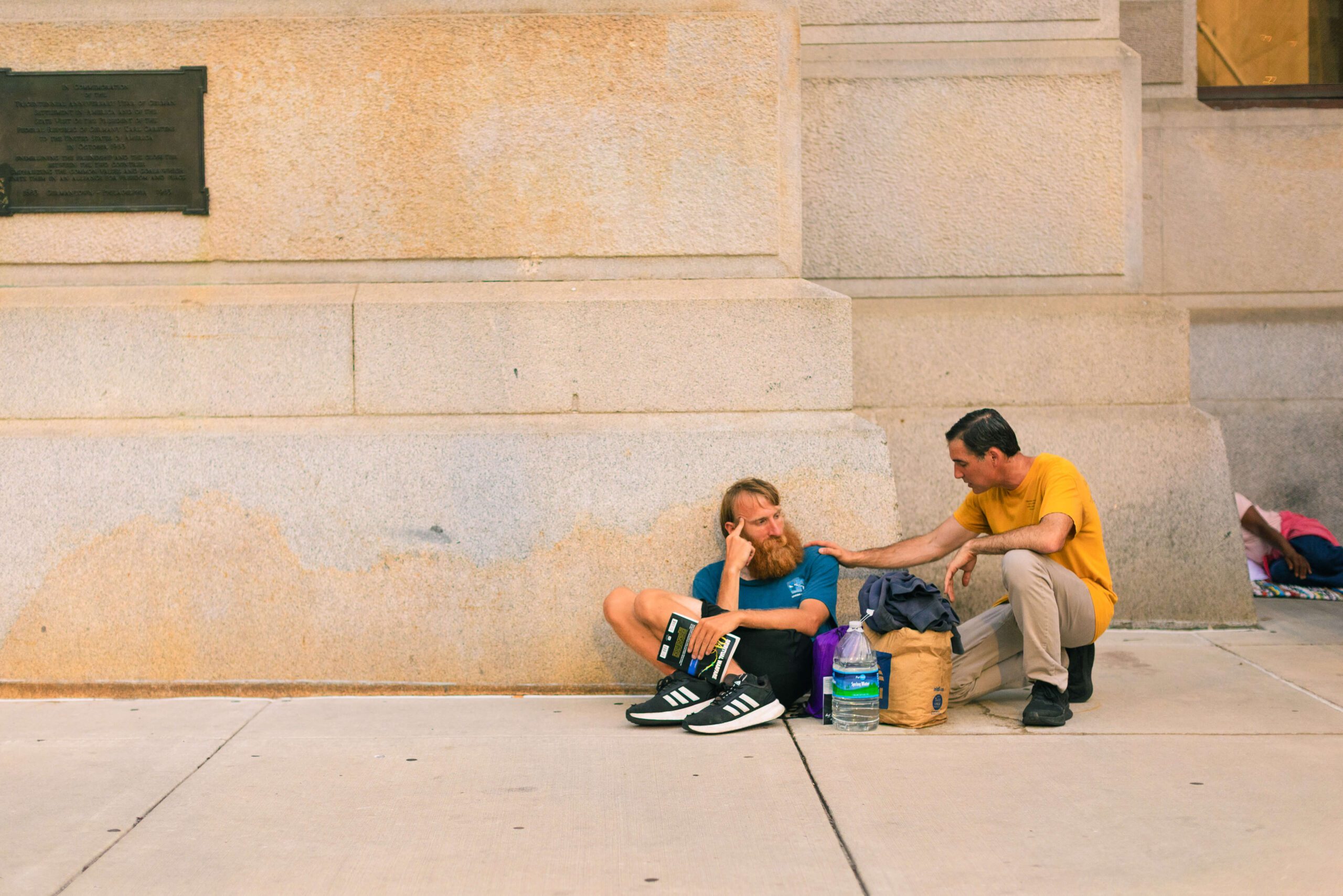 A man with a beard sitting on the sidewalk appears contemplative while wearing a blue shirt and black sneakers. Another man in a yellow shirt is kneeling beside him, offering support. They are near a stone wall, with bags and a water bottle visible nearby, capturing a moment of compassion in an urban setting.