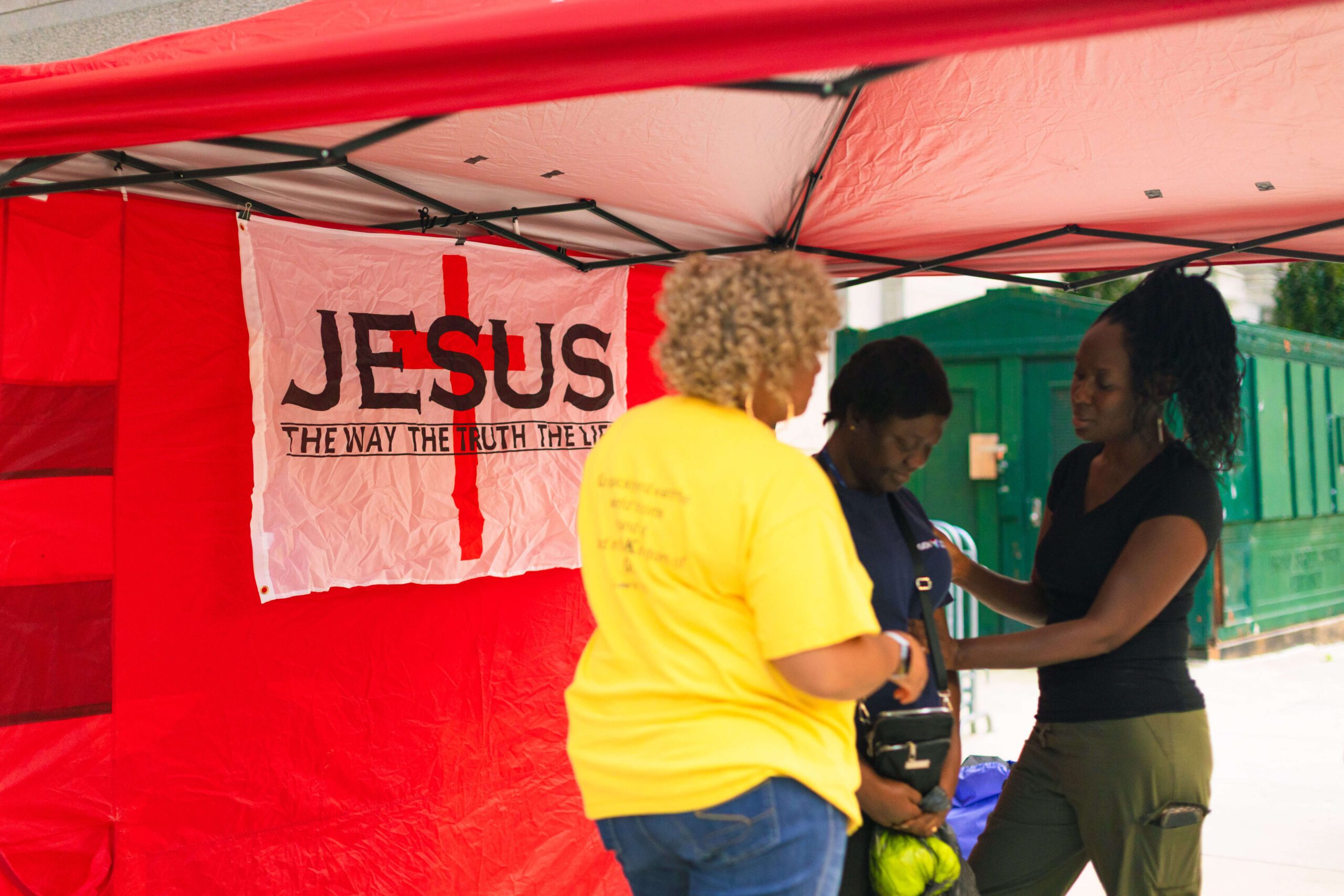 Two women interact under a red tent featuring a banner that reads "JESUS" with the phrase "THE WAY THE TRUTH THE LIFE." One woman in a yellow shirt is speaking to another who is wearing a blue shirt, while a third woman in black helps them. The setting appears to be a community outreach event.